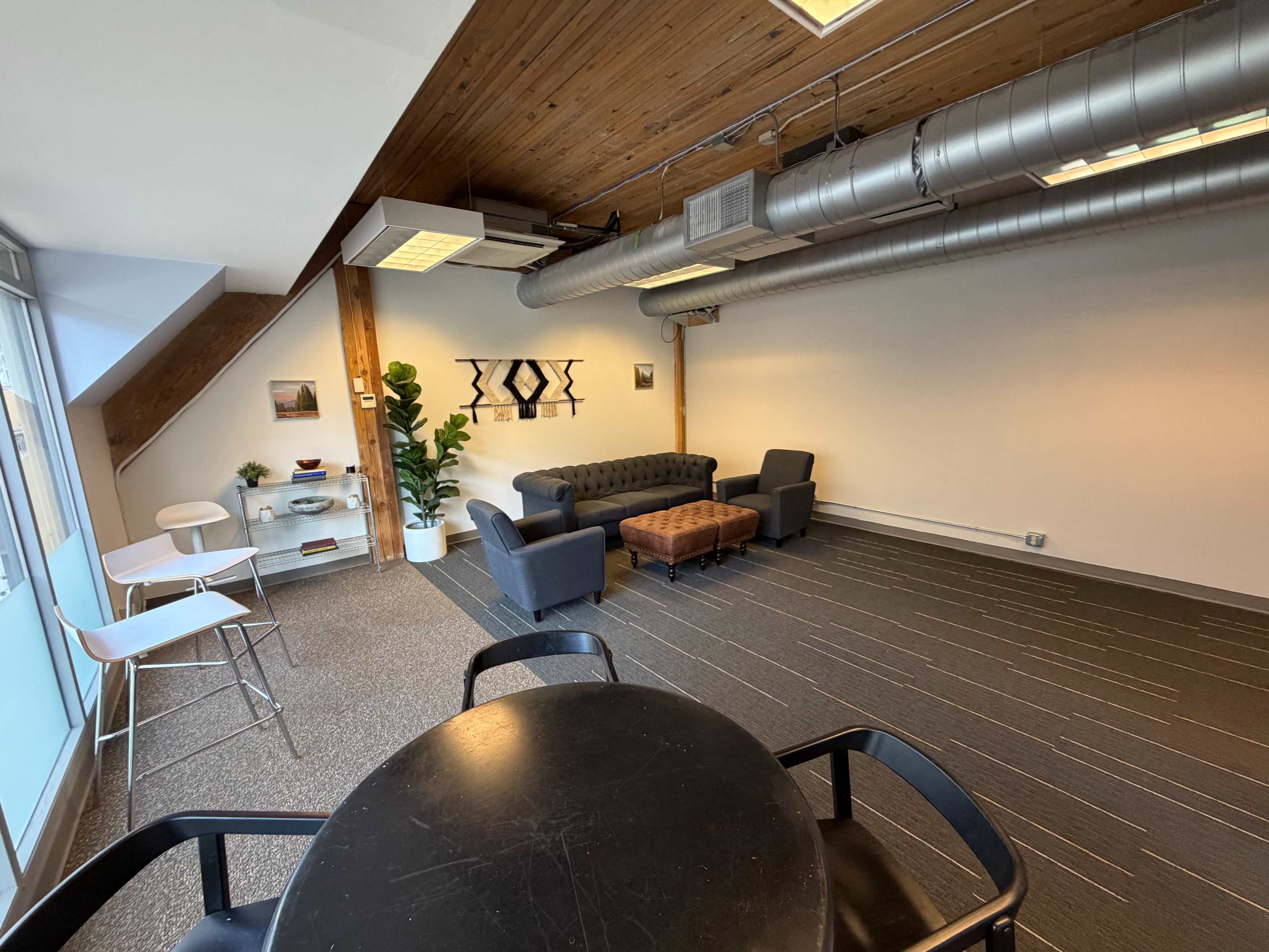 The image shows a spacious, modern office/lounge area featuring a black couch, a coffee table, two chairs, and large windows that provide natural light.