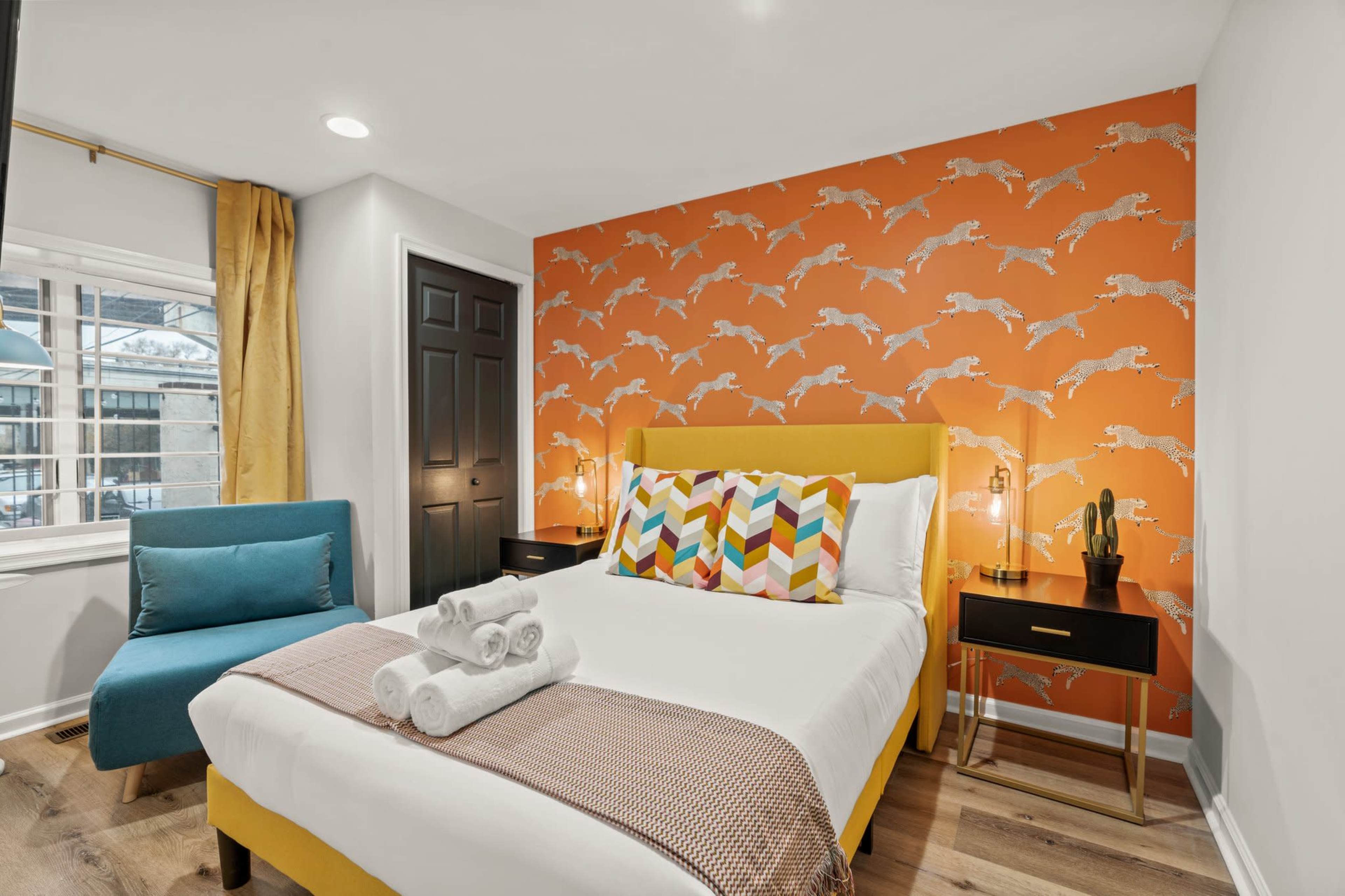 The image shows a brightly decorated bedroom with a yellow bed, colorful pillows, an orange patterned accent wall, and a blue armchair in the corner.