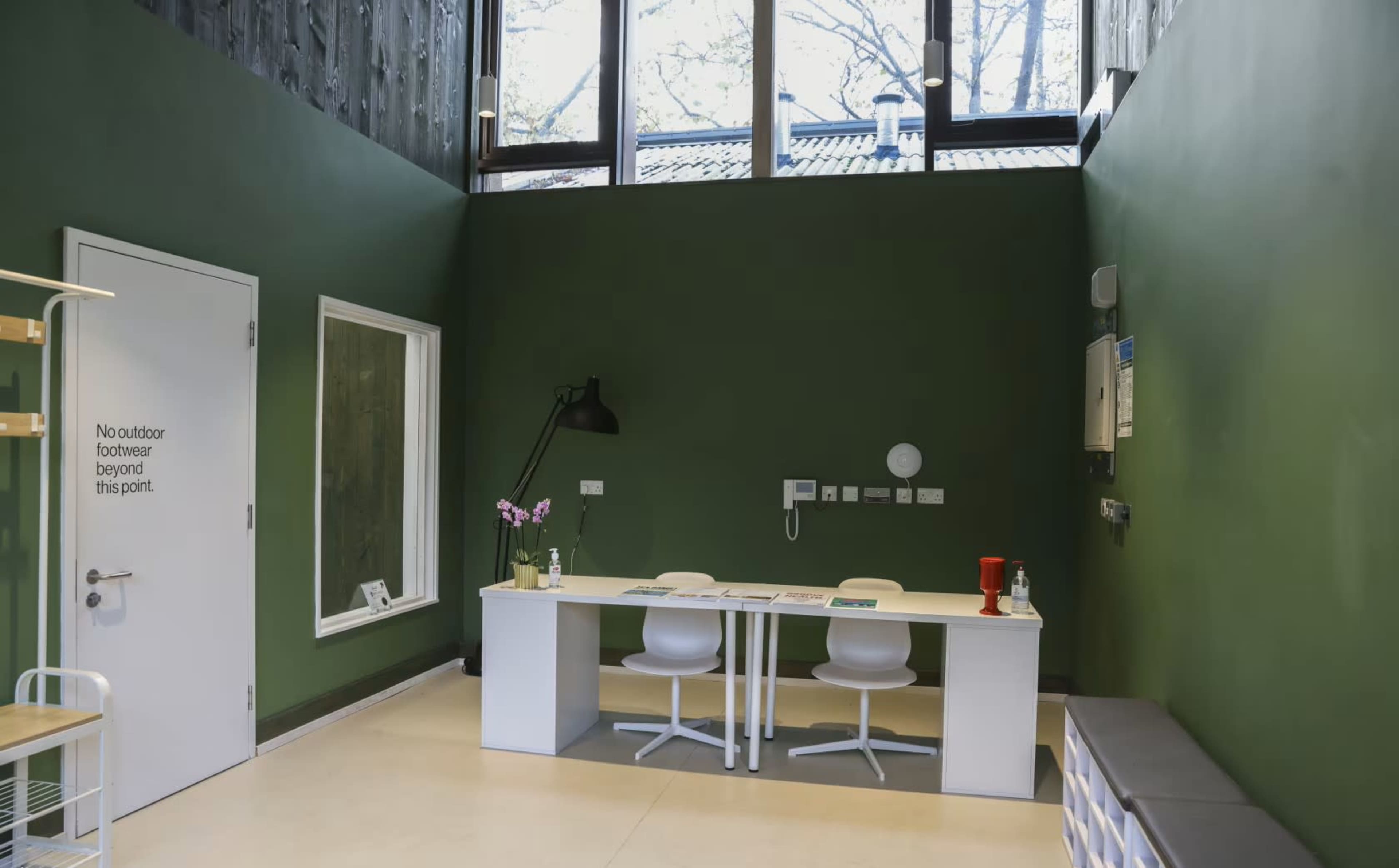 The image shows a modern office space with a green wall, a white table, two chairs, and large windows allowing natural light to enter.
