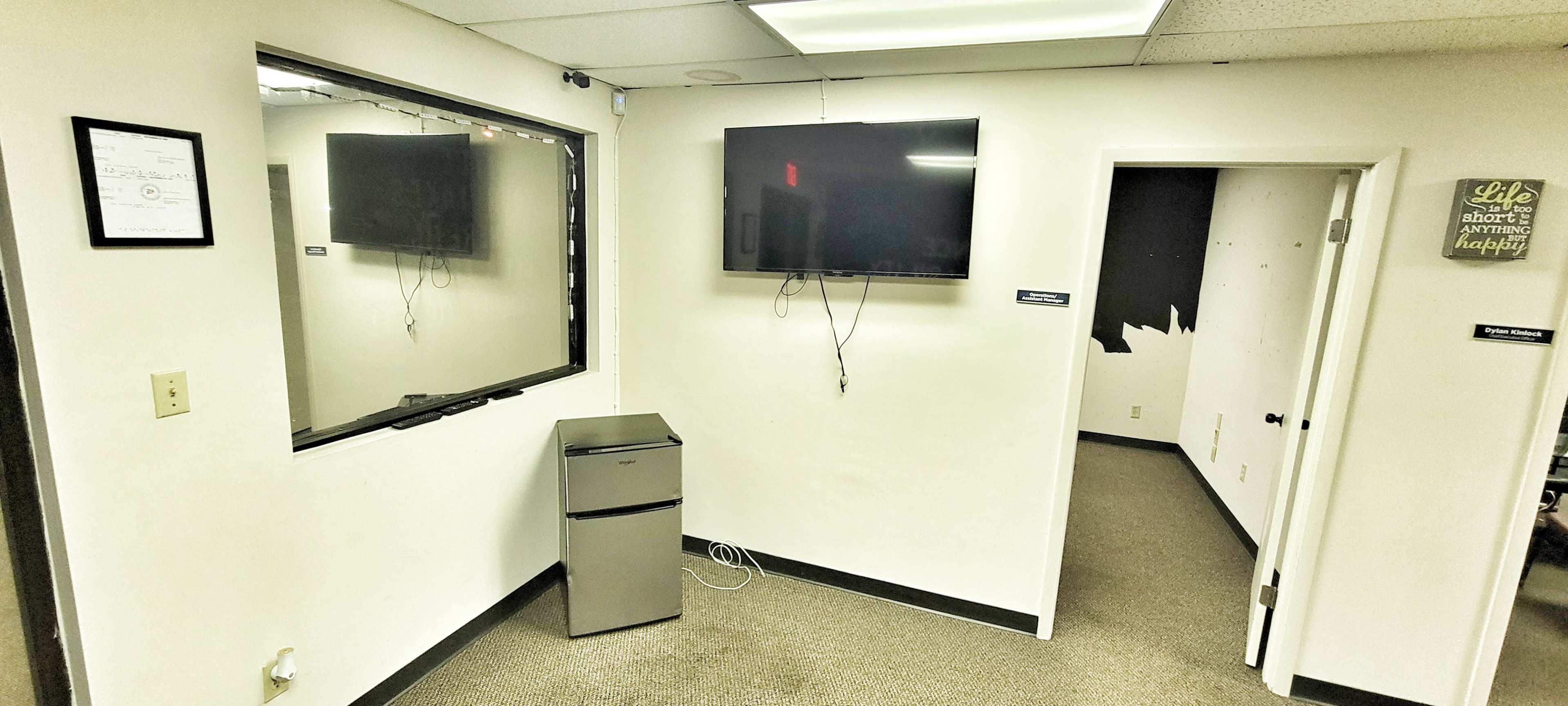 The image shows a small room with a mirror, a mounted television on the wall, a mini-fridge, and a door leading to another area.