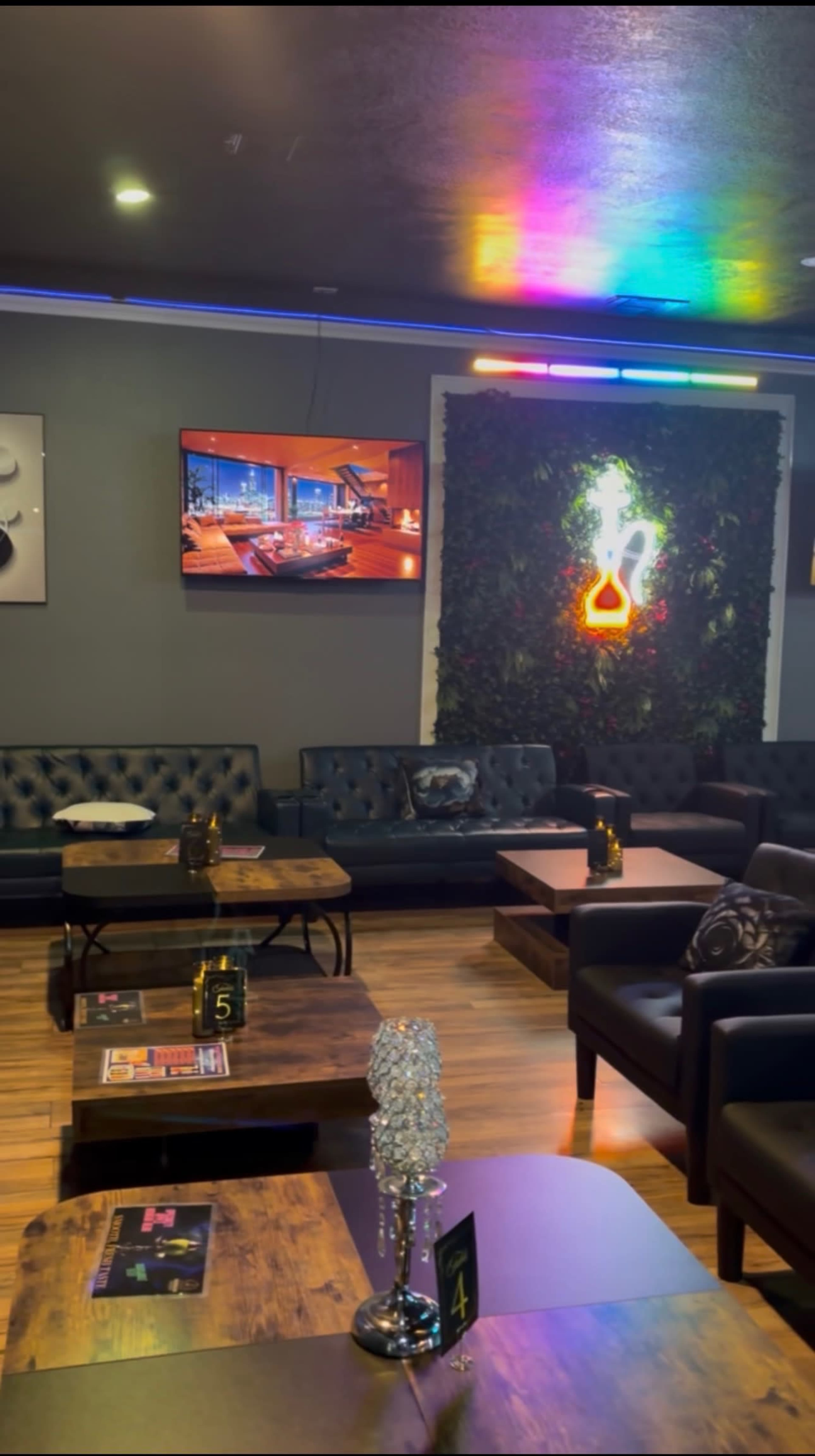 The image shows a modern lounge area with dark sofas, wooden tables, a neon decoration, and a television on the wall displaying a cozy interior scene.
