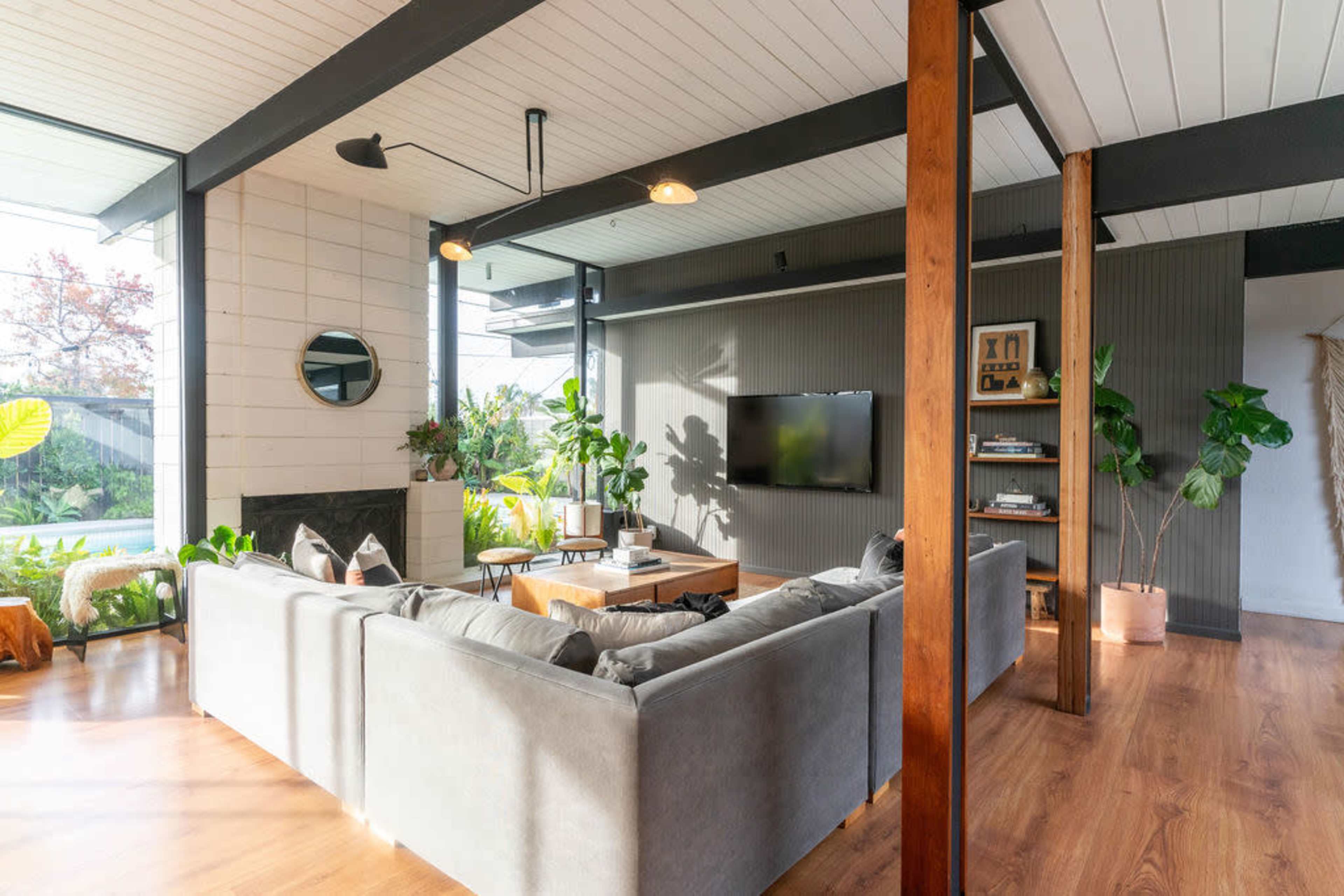 The image depicts a modern living room featuring a large gray sectional sofa, wooden accents, potted plants, and a television mounted on the wall.