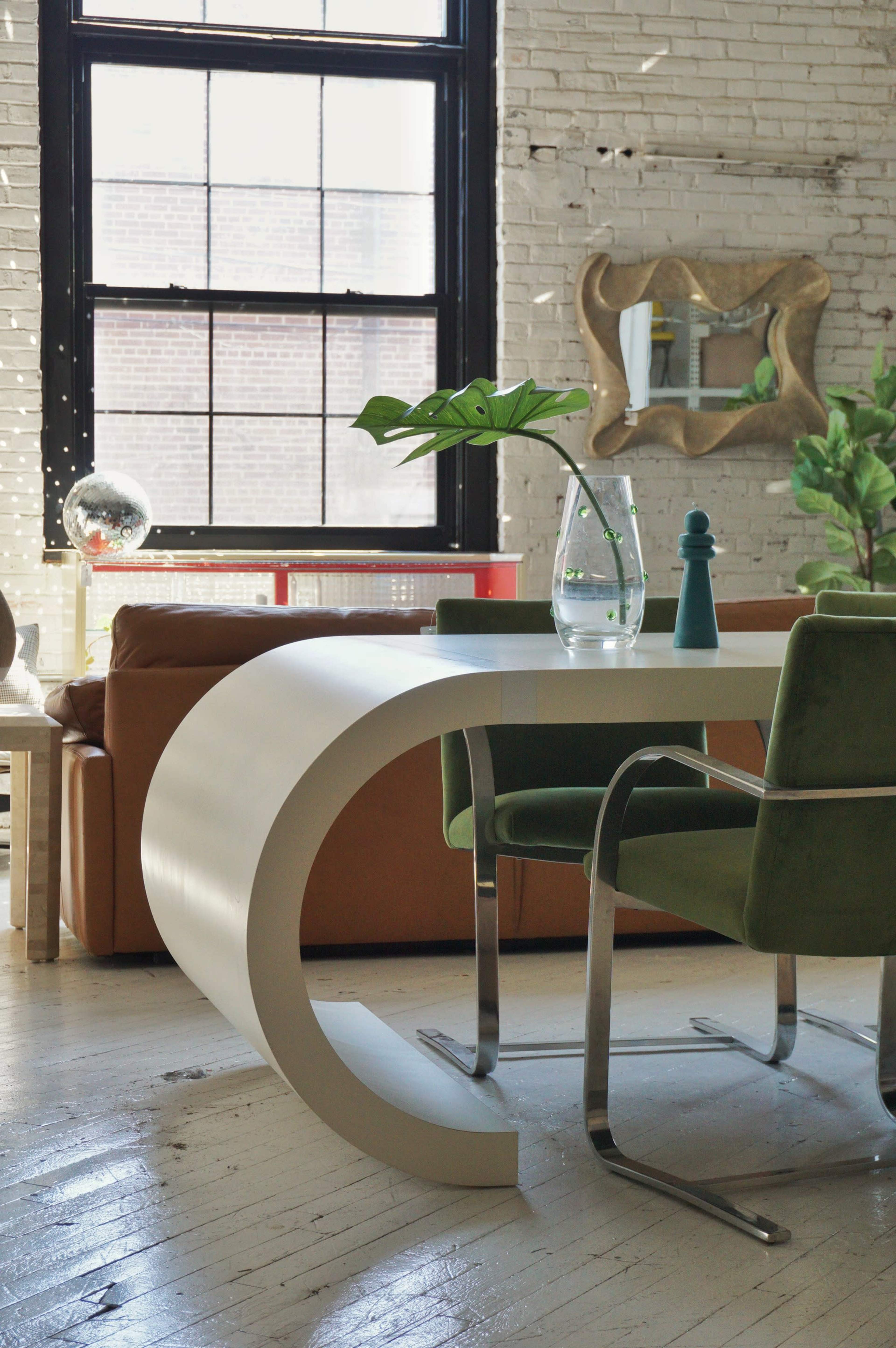 The image shows a modern, curved white desk paired with green upholstered chairs in a bright room with large windows and minimalist decor.