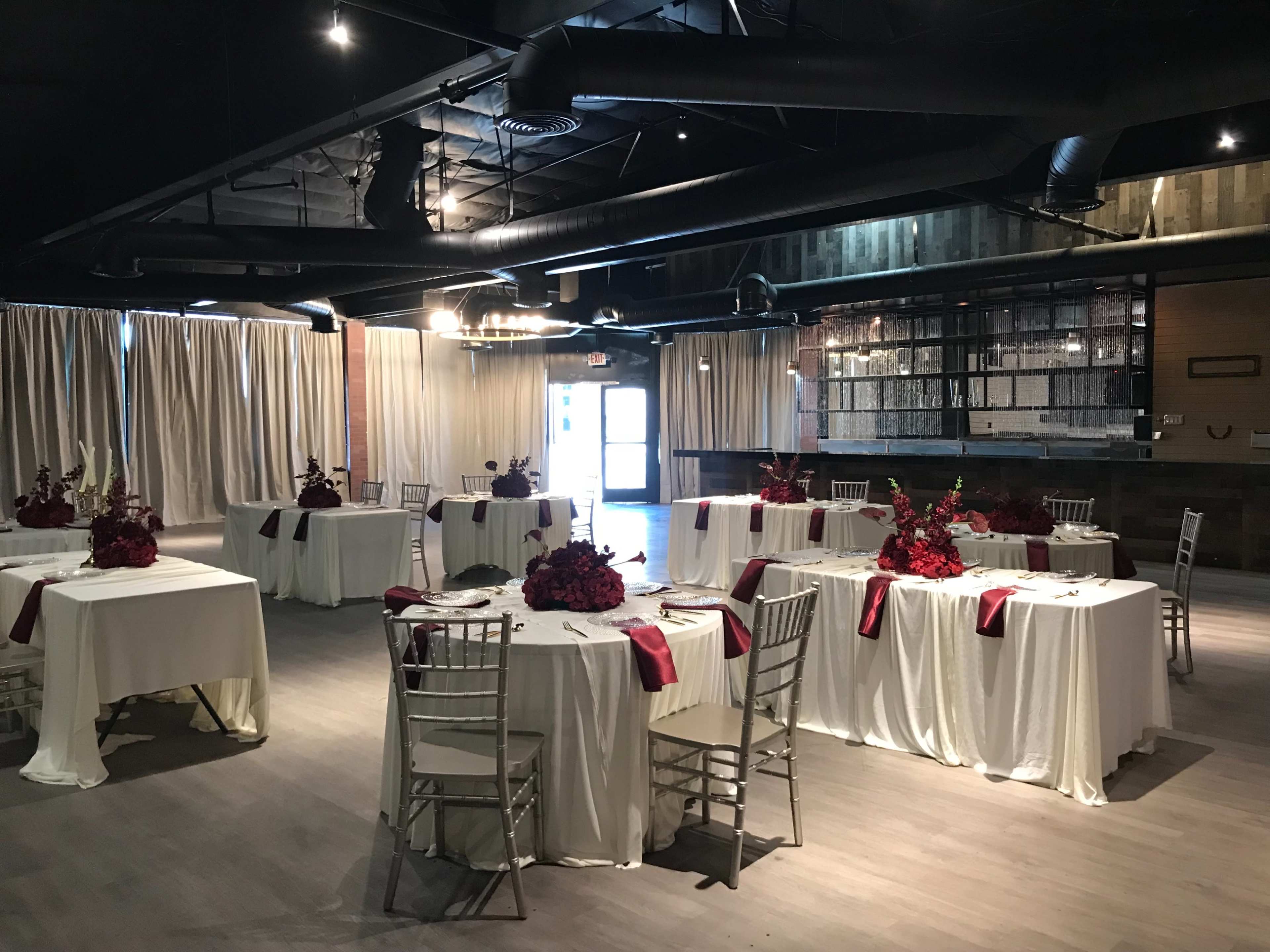 The image shows a banquet hall set up with several tables adorned with white tablecloths and red floral centerpieces.