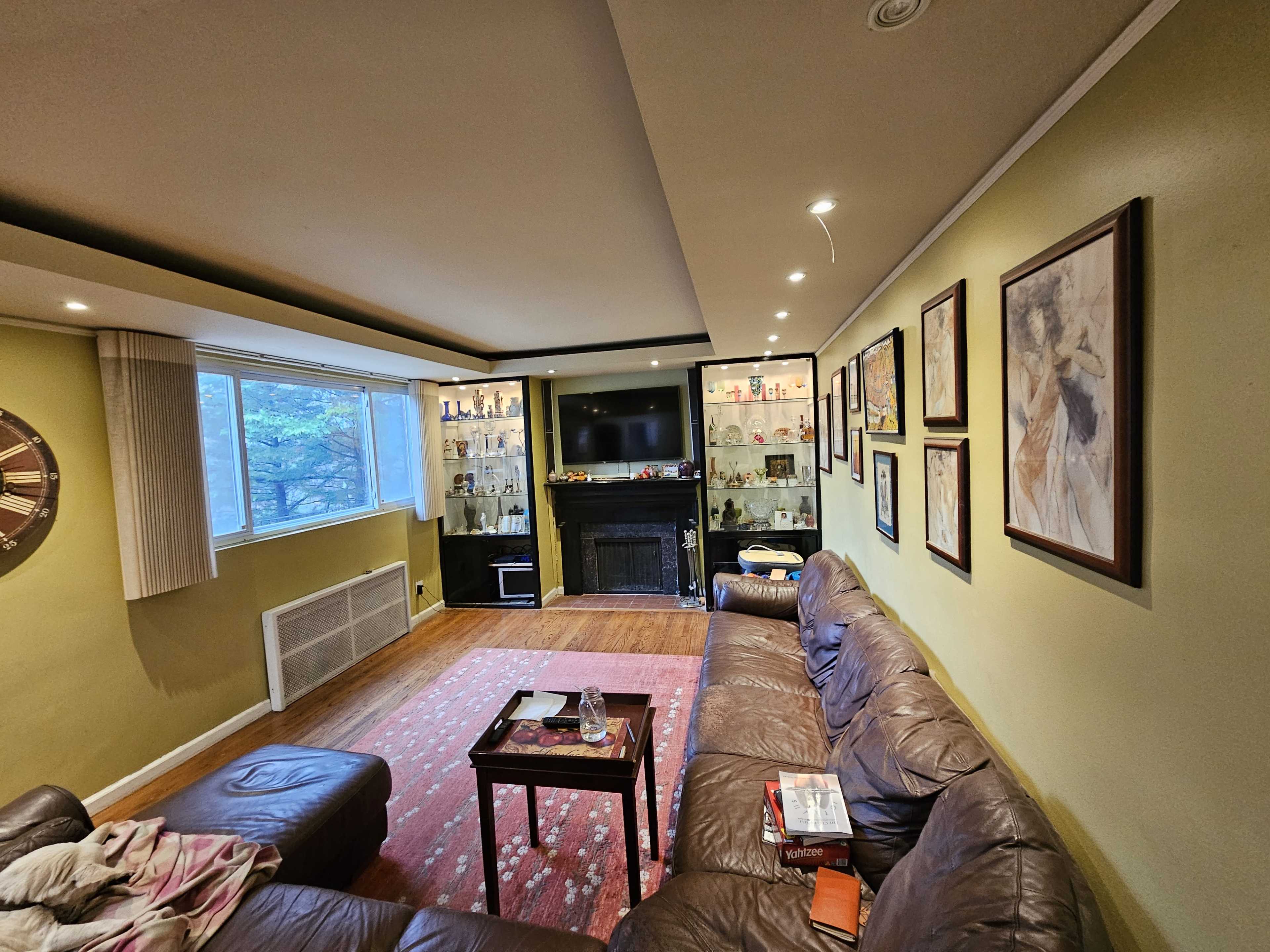 A living room with a brown leather sectional couch, a coffee table, and a wall decorated with framed art, alongside shelves displaying various items and a television mounted above a fireplace.