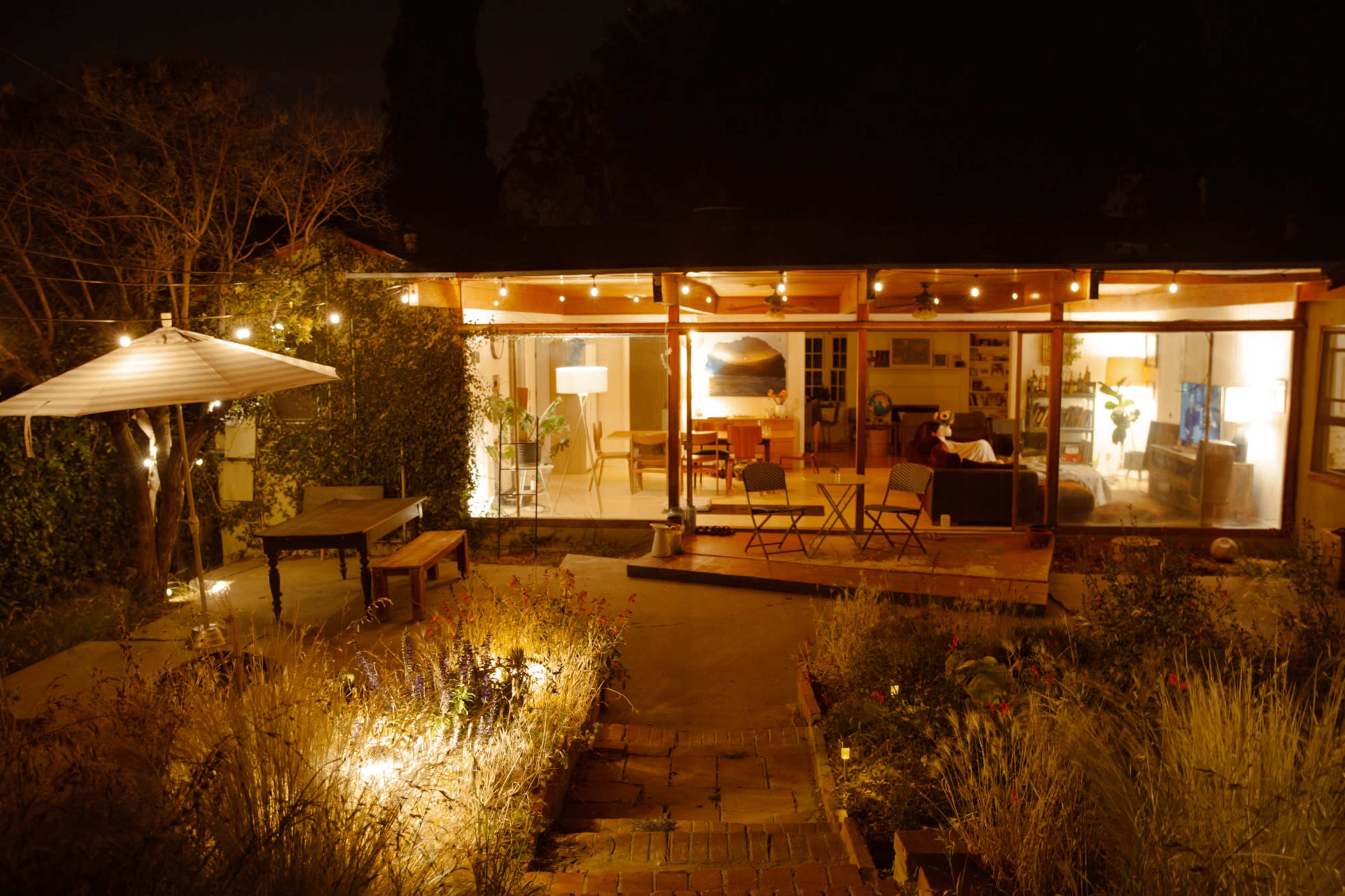A well-lit modern house with large glass windows is surrounded by a garden at night, featuring outdoor seating and string lights.