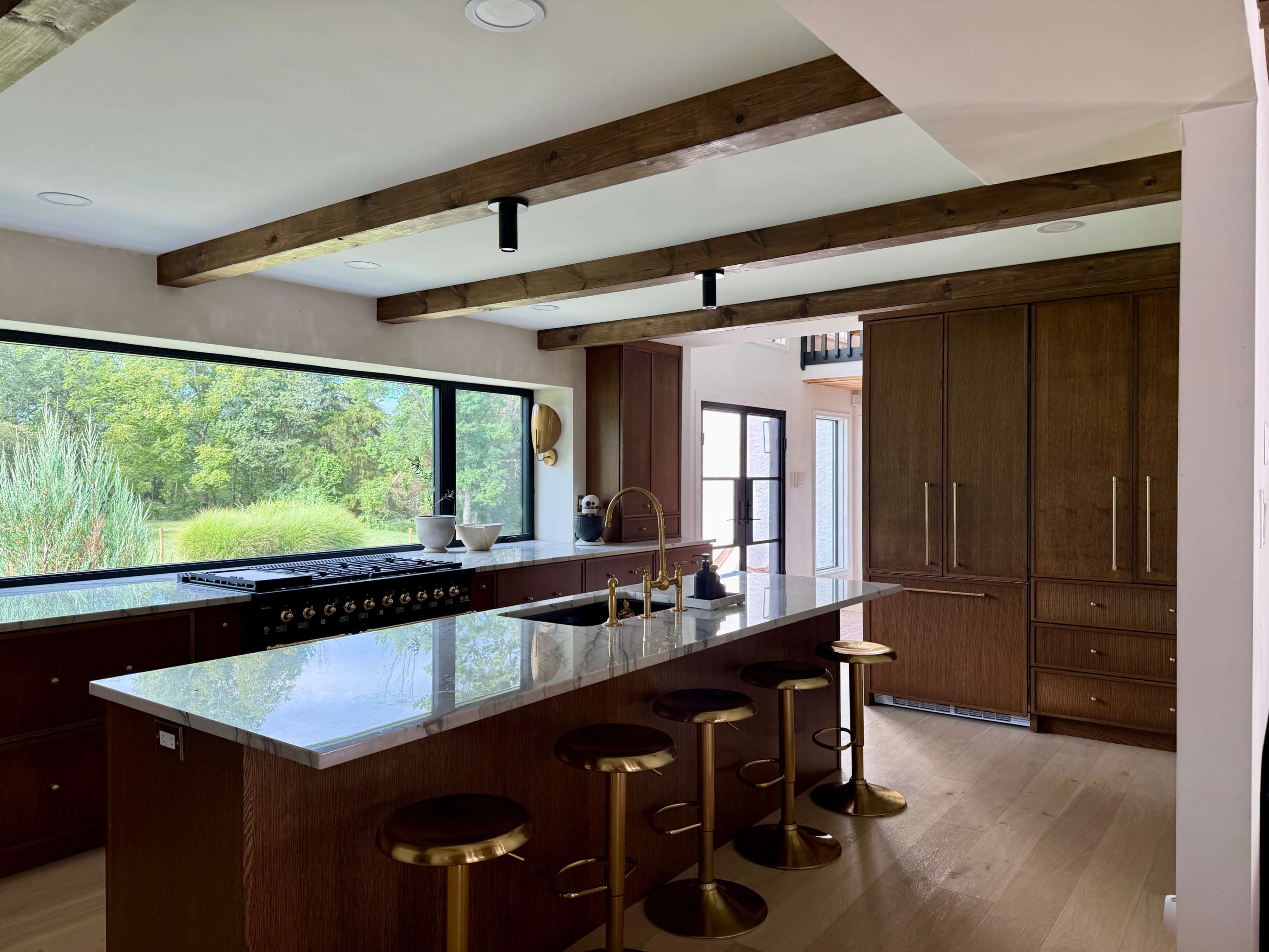 The image shows a modern kitchen featuring a large island with bar stools, wooden beams on the ceiling, and a panoramic window overlooking a green landscape.