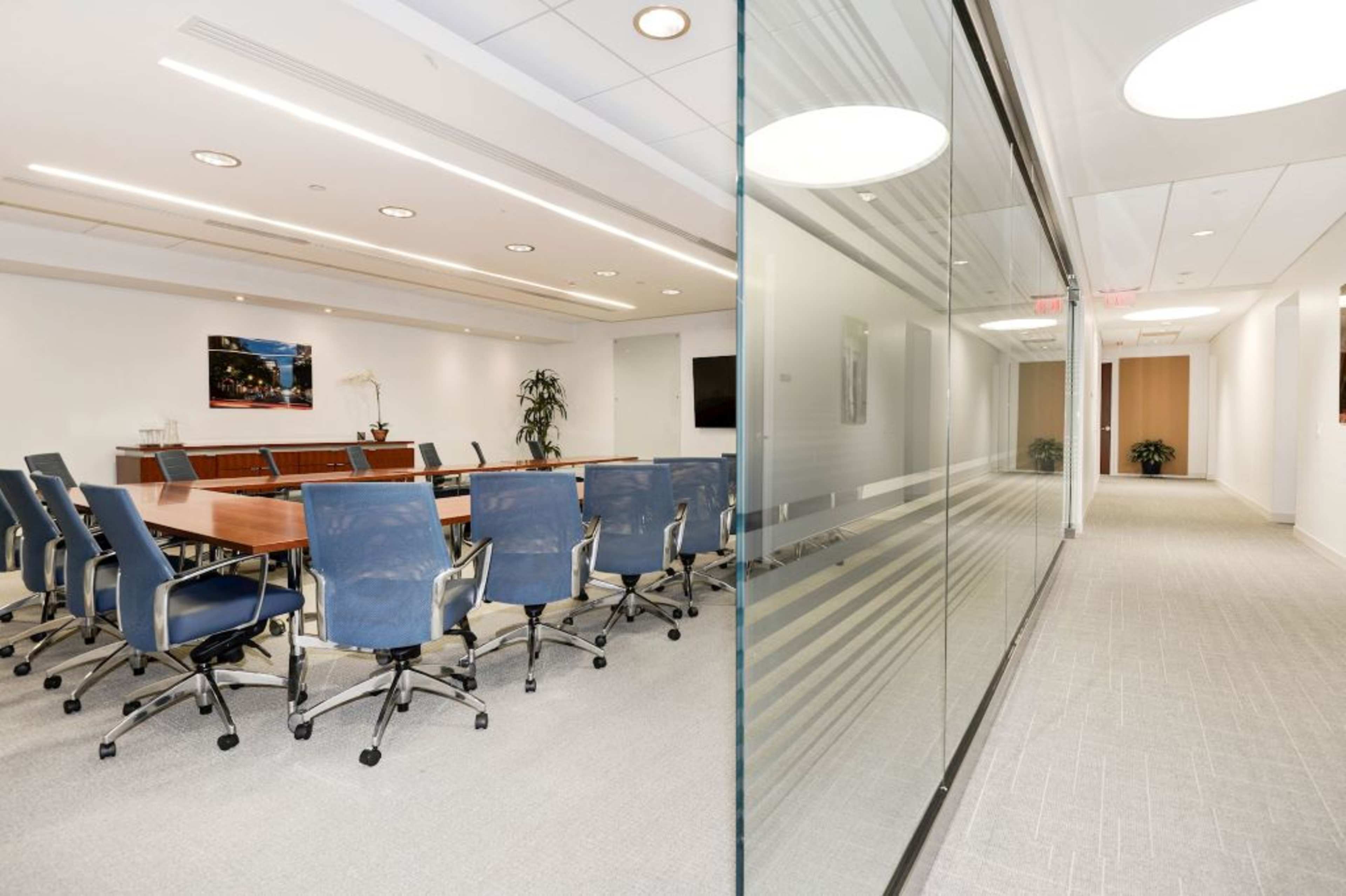 A modern conference room with a large wooden table surrounded by blue chairs, visible through a glass partition leading to a hallway.