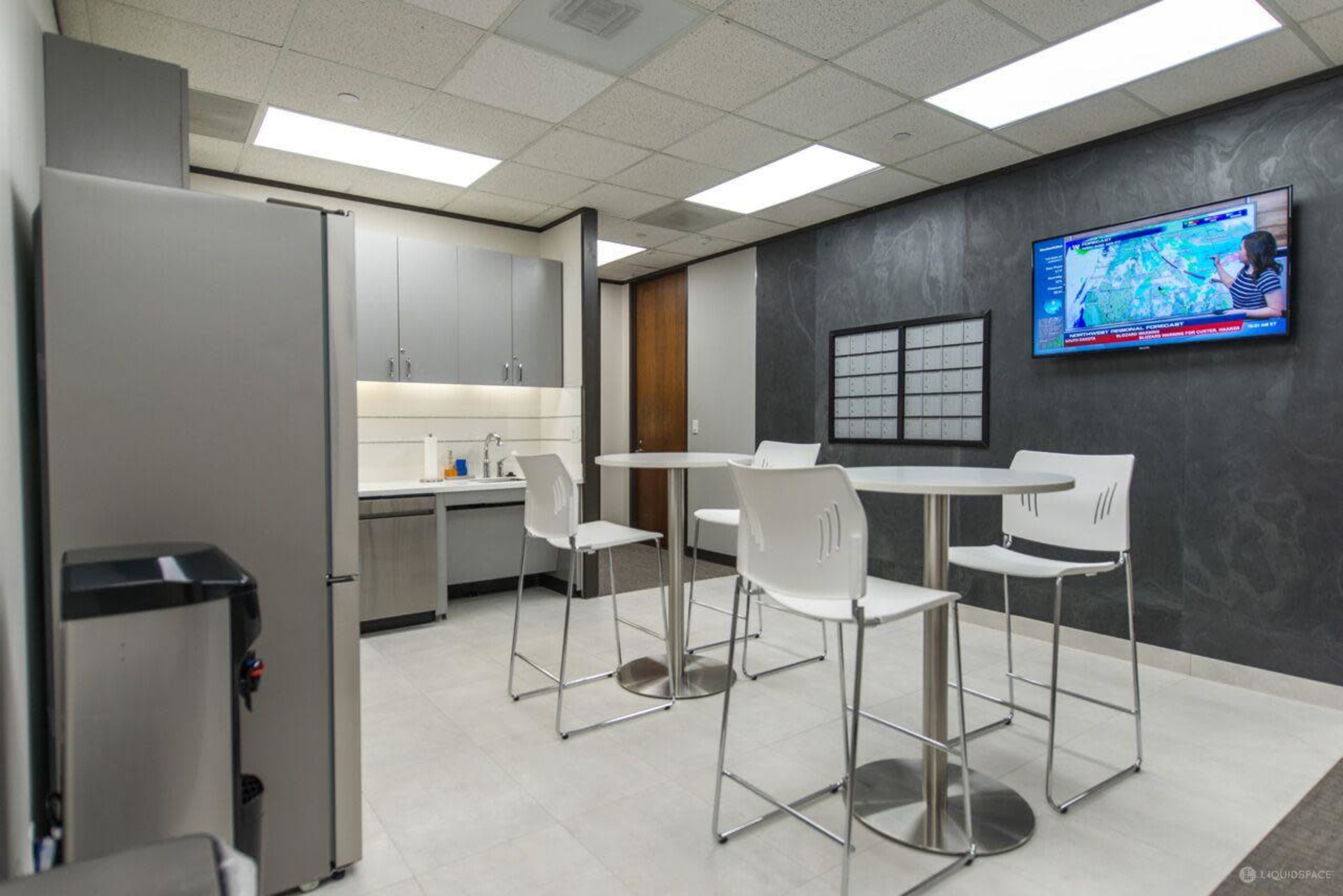 A modern break room features a kitchenette with a refrigerator, a large table with four high chairs, and a television mounted on the wall displaying news.