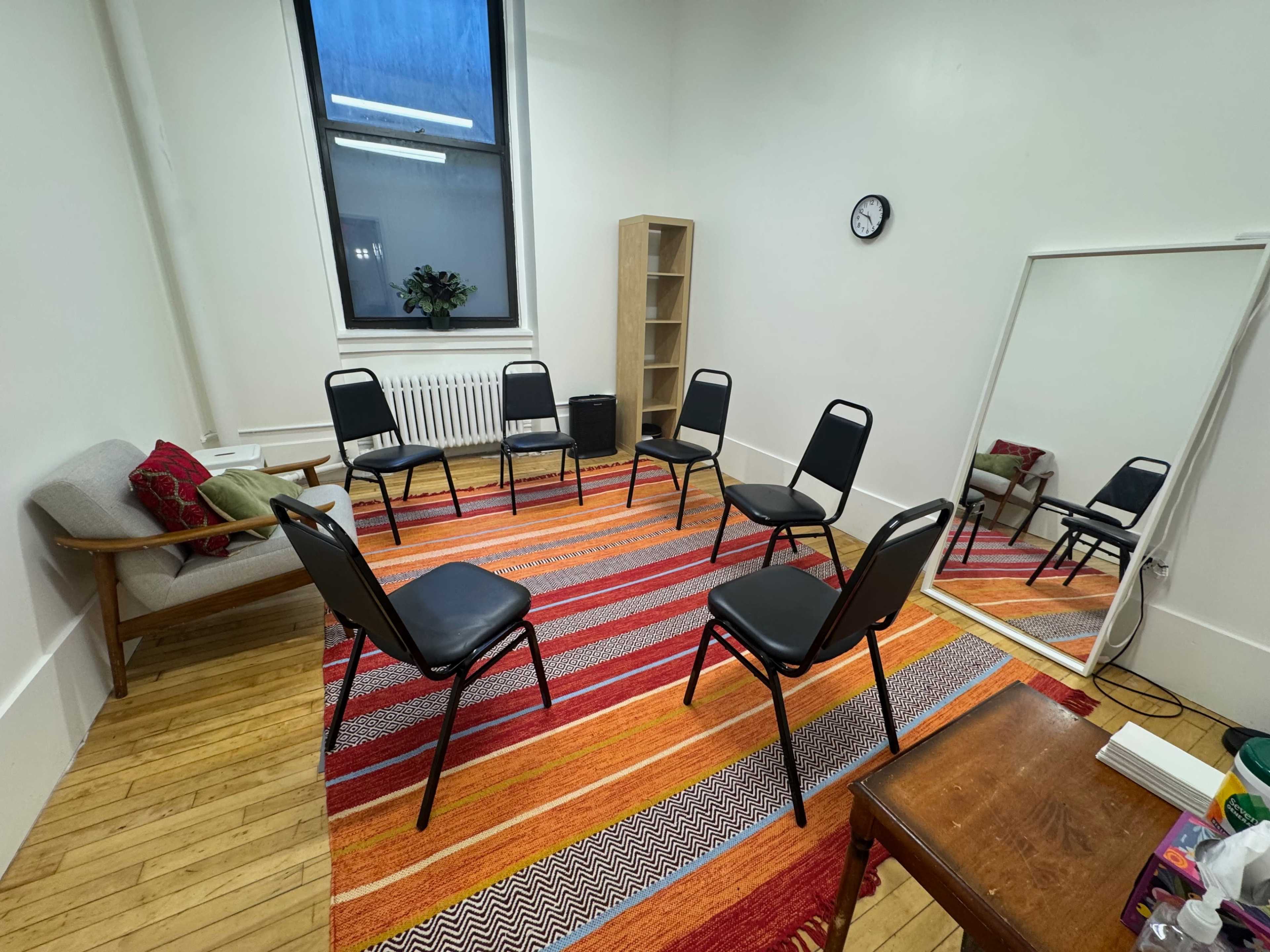 The image shows a small meeting room with eight black chairs arranged in a circle on a colorful striped rug, along with a bookshelf, mirror, and a clock on the wall.