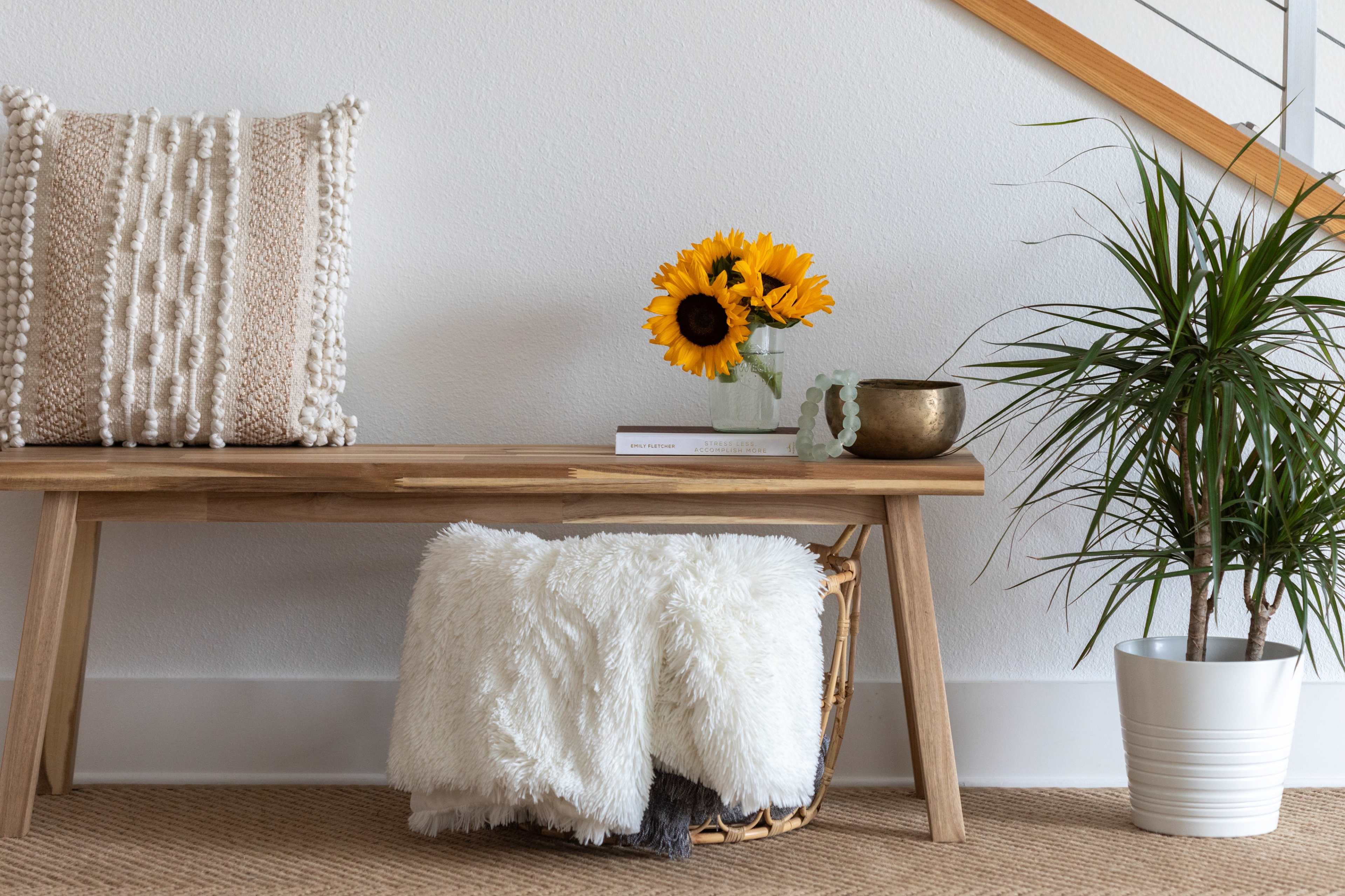 A wooden bench with a decorative pillow holds a vase of sunflowers and a bowl, beside a potted plant and a cozy throw blanket in a woven basket.