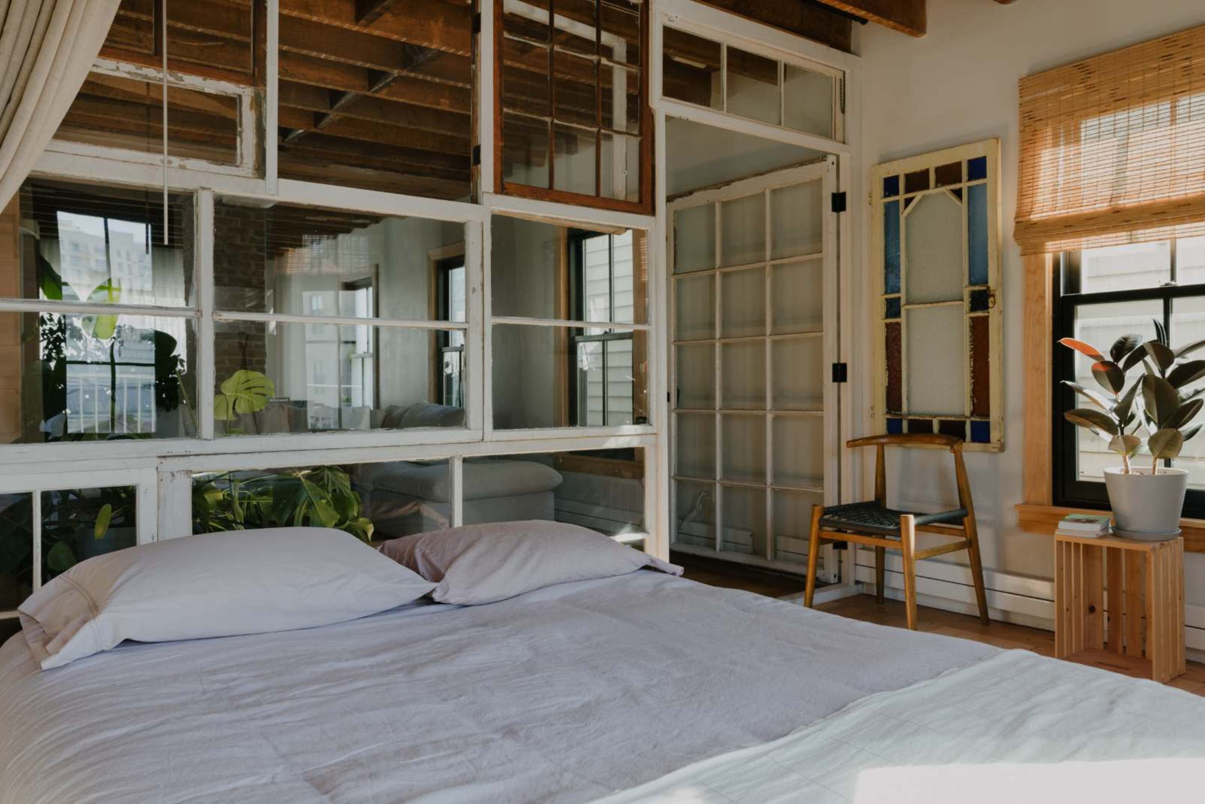 A bedroom features a bed with two pillows, surrounded by large windows that allow natural light to illuminate the space and showcase a seating area with a chair and plants.