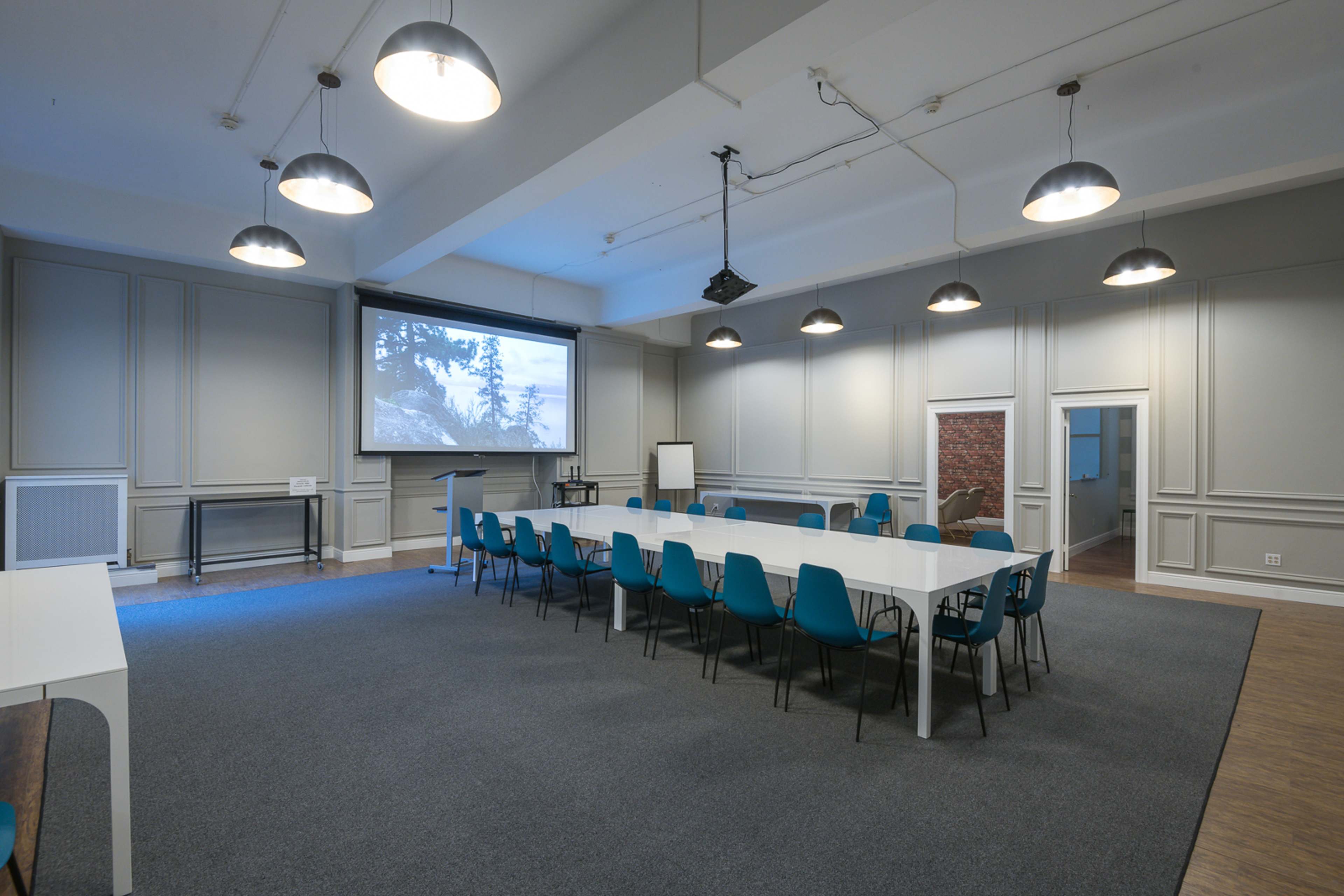 A spacious meeting room features a long white table surrounded by turquoise chairs, with a projection screen at the front and large windows letting in natural light.