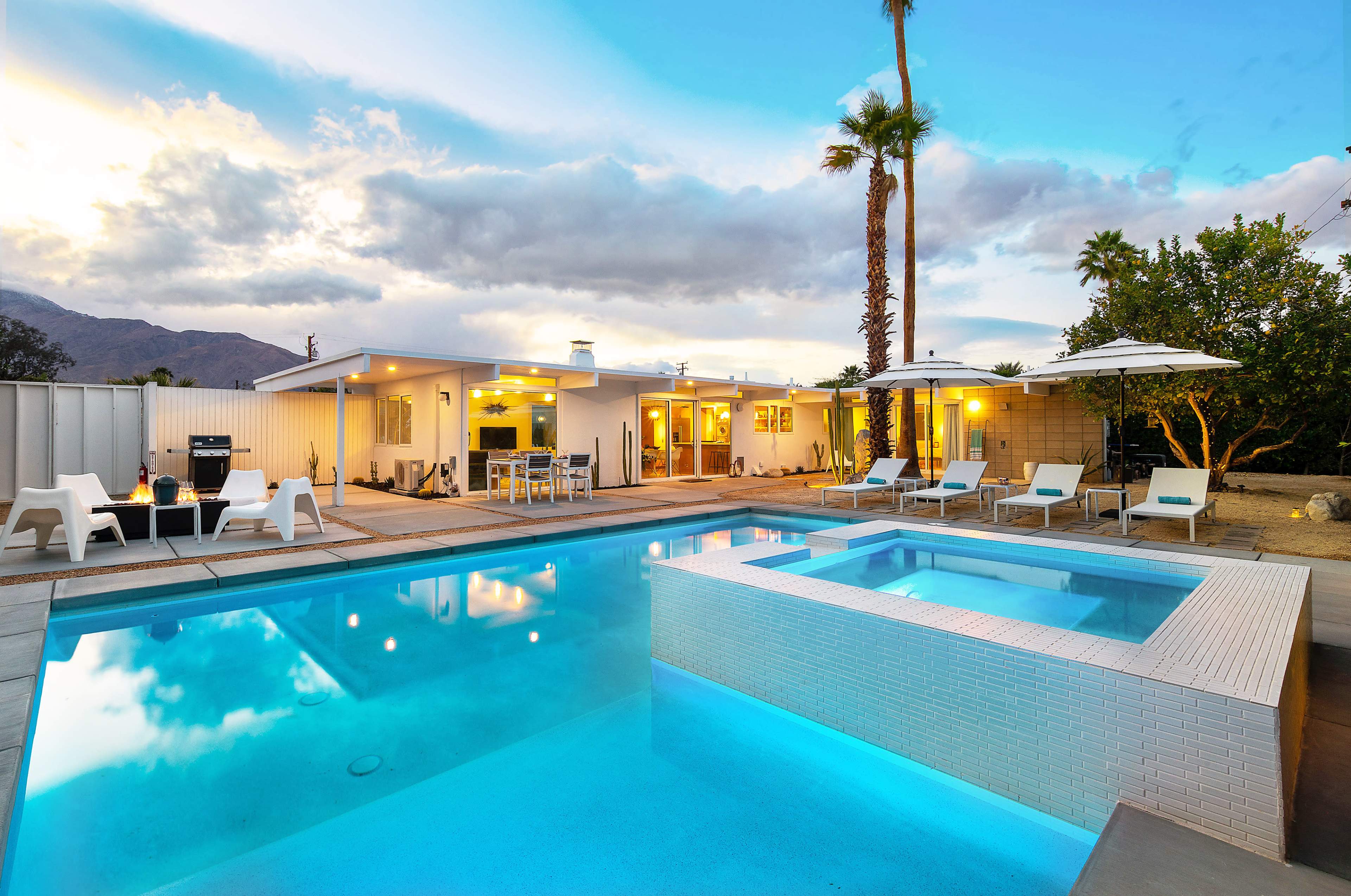 The scene features a modern swimming pool with a jacuzzi surrounded by lounge chairs, palm trees, and a single-story house under a cloudy sky.