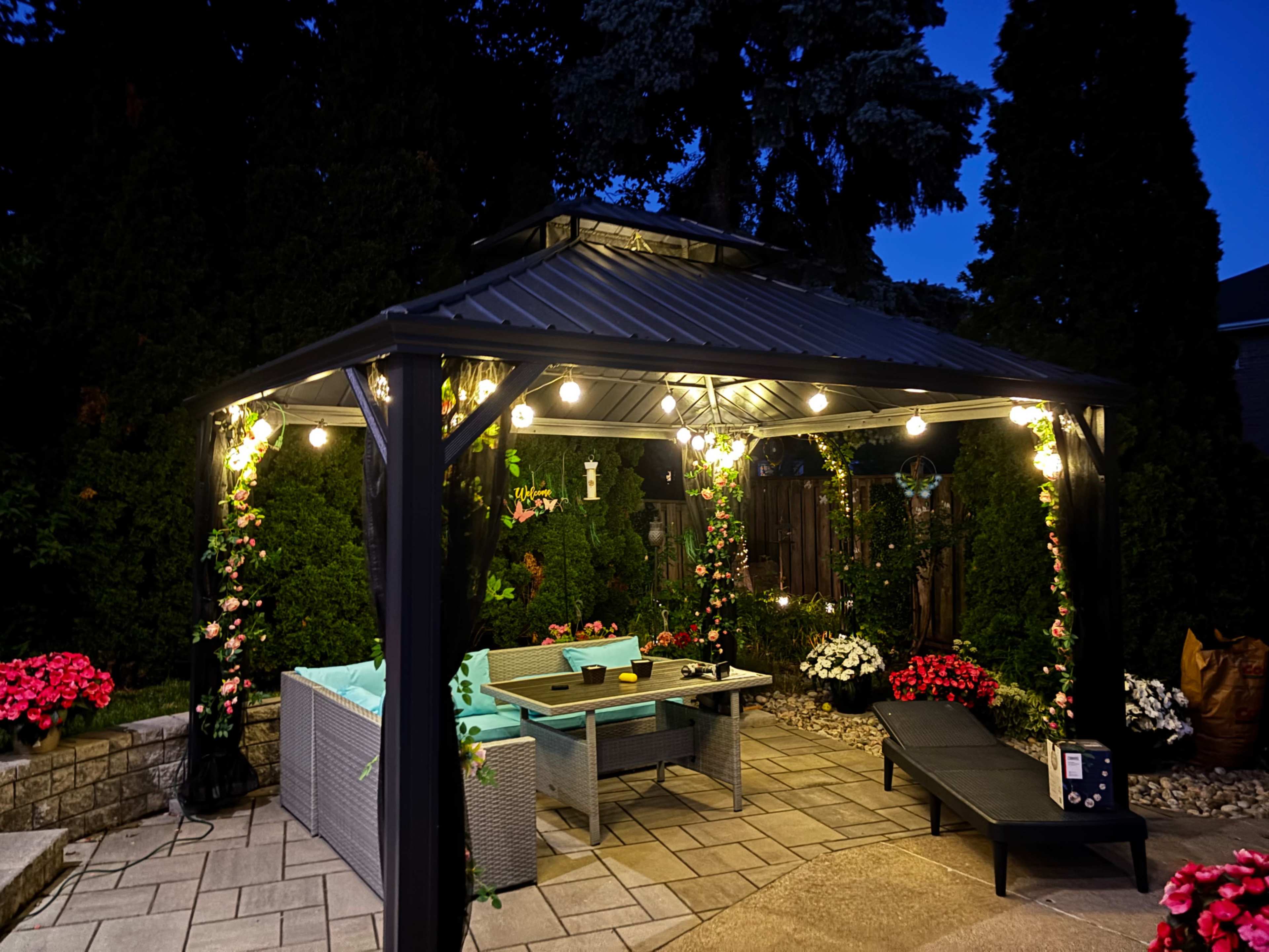 A garden gazebo with string lights is surrounded by flower beds and patio furniture, set against a twilight sky.
