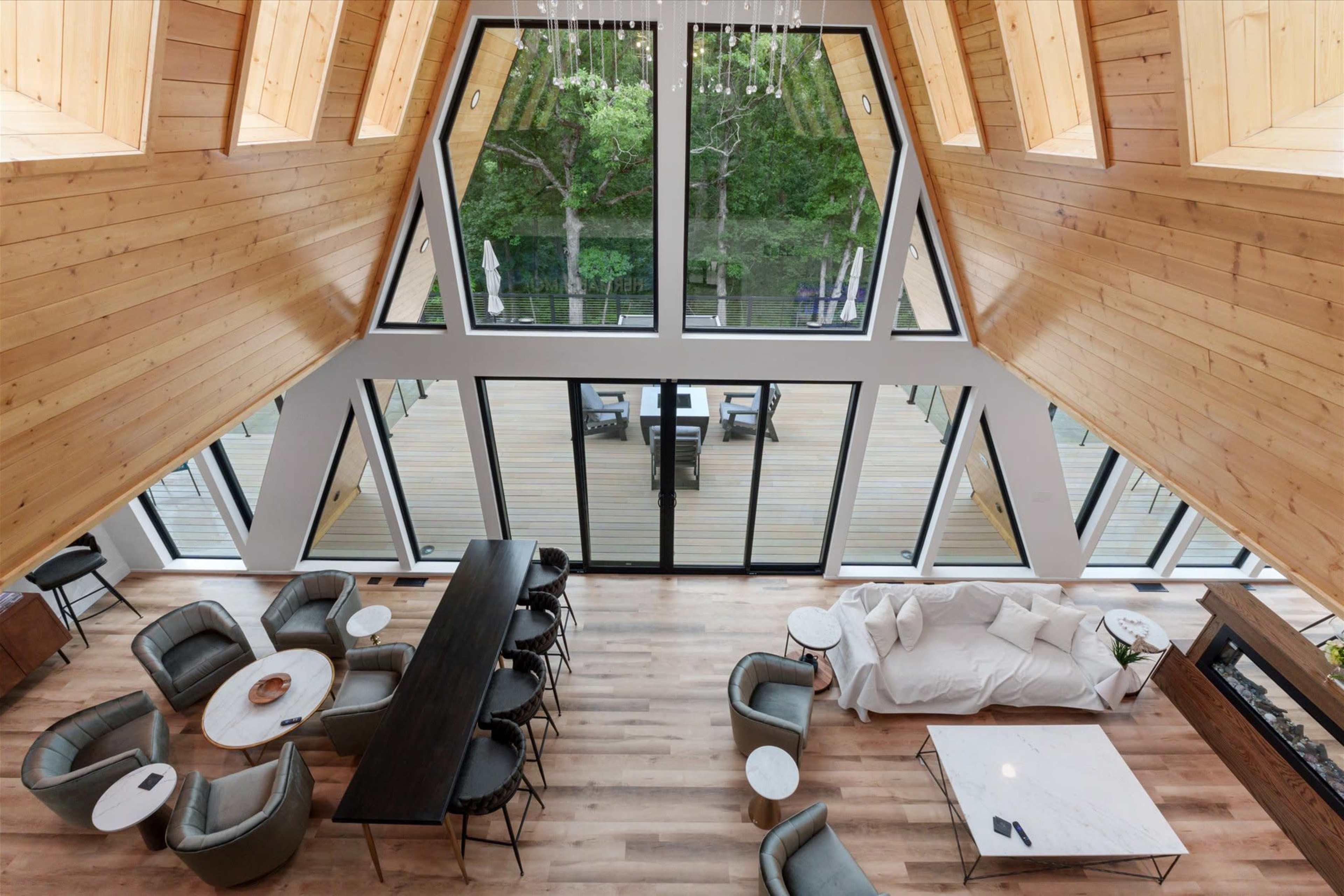 The image shows a modern interior of an A-frame house featuring large glass windows, wooden walls, a dining area with a long table, and a comfortable seating arrangement.