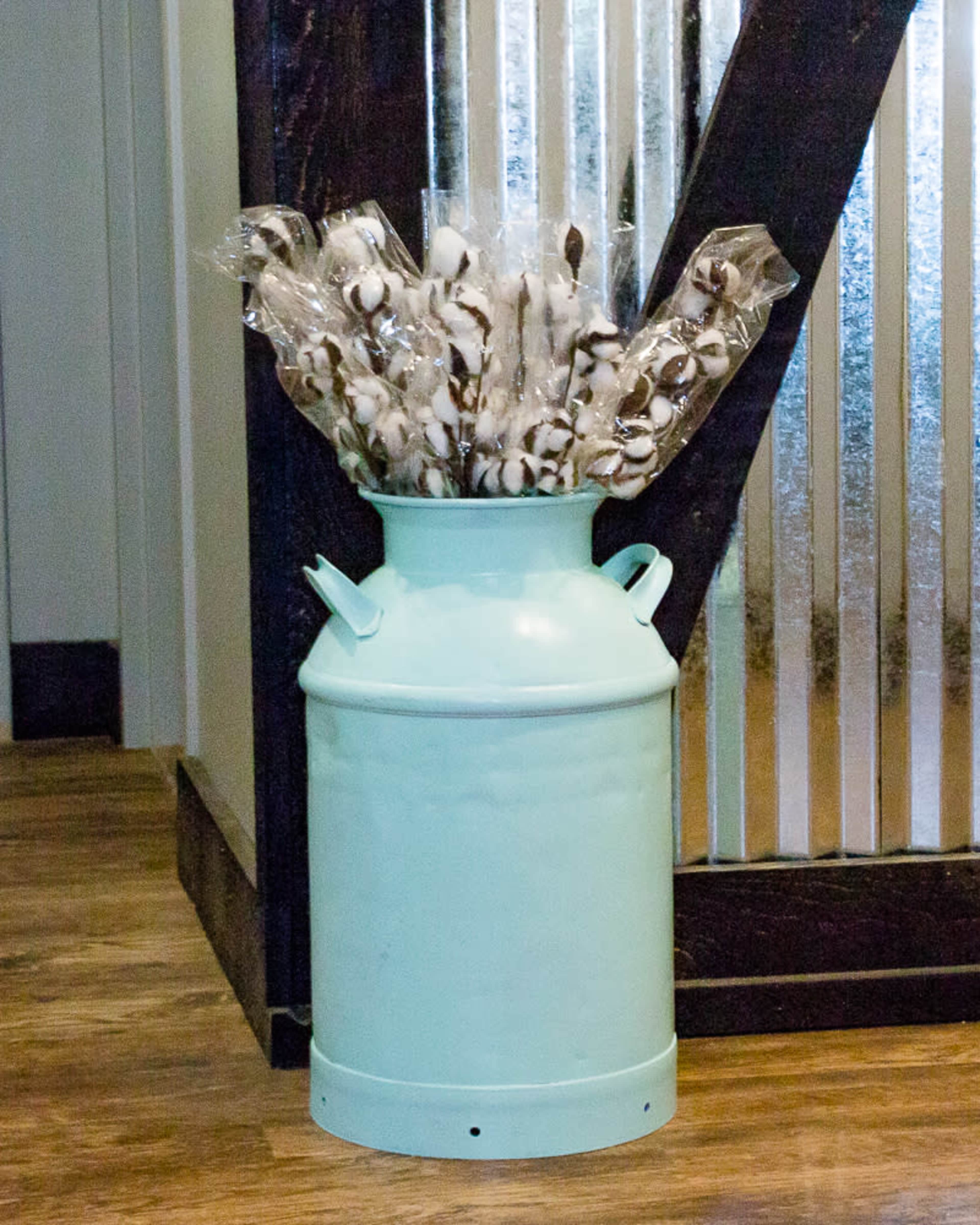 A turquoise metal milk can holds a bundle of cotton stems wrapped in plastic and placed against a textured wall.