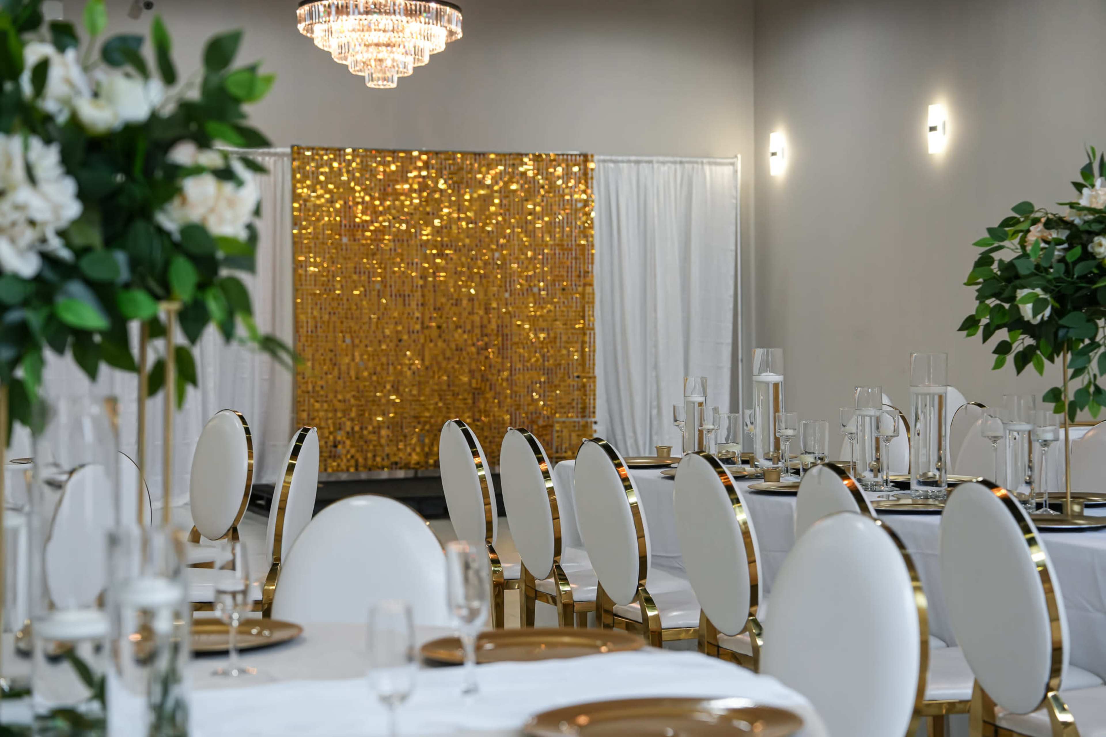 A decorated dining area with round tables set for an event, featuring white tablecloths, gold accents, and a shimmering golden backdrop.