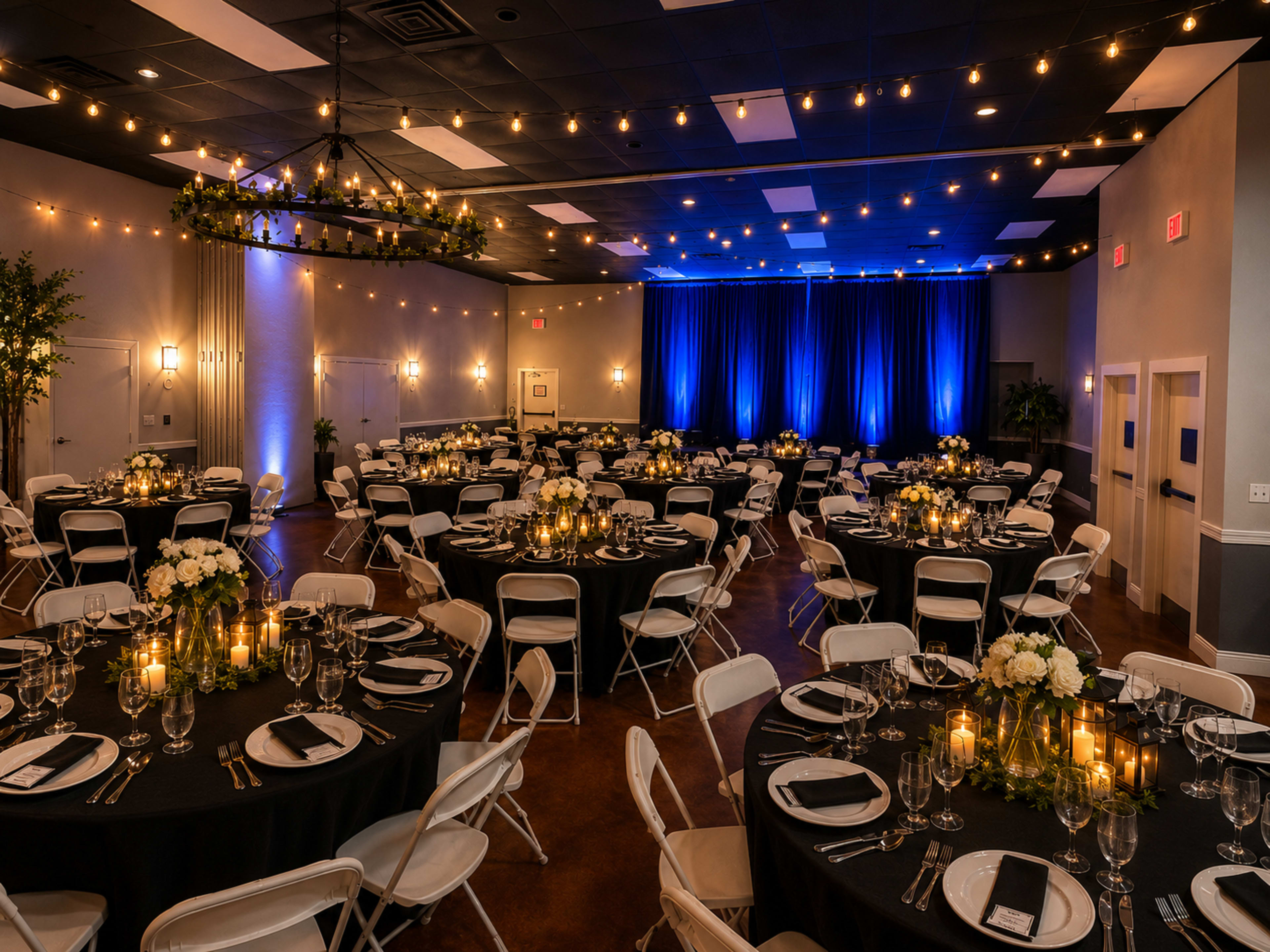 The image shows a banquet hall set up for an event, featuring round tables with black tablecloths, white chairs, and softly lit candles, along with a stage area illuminated by blue lighting.