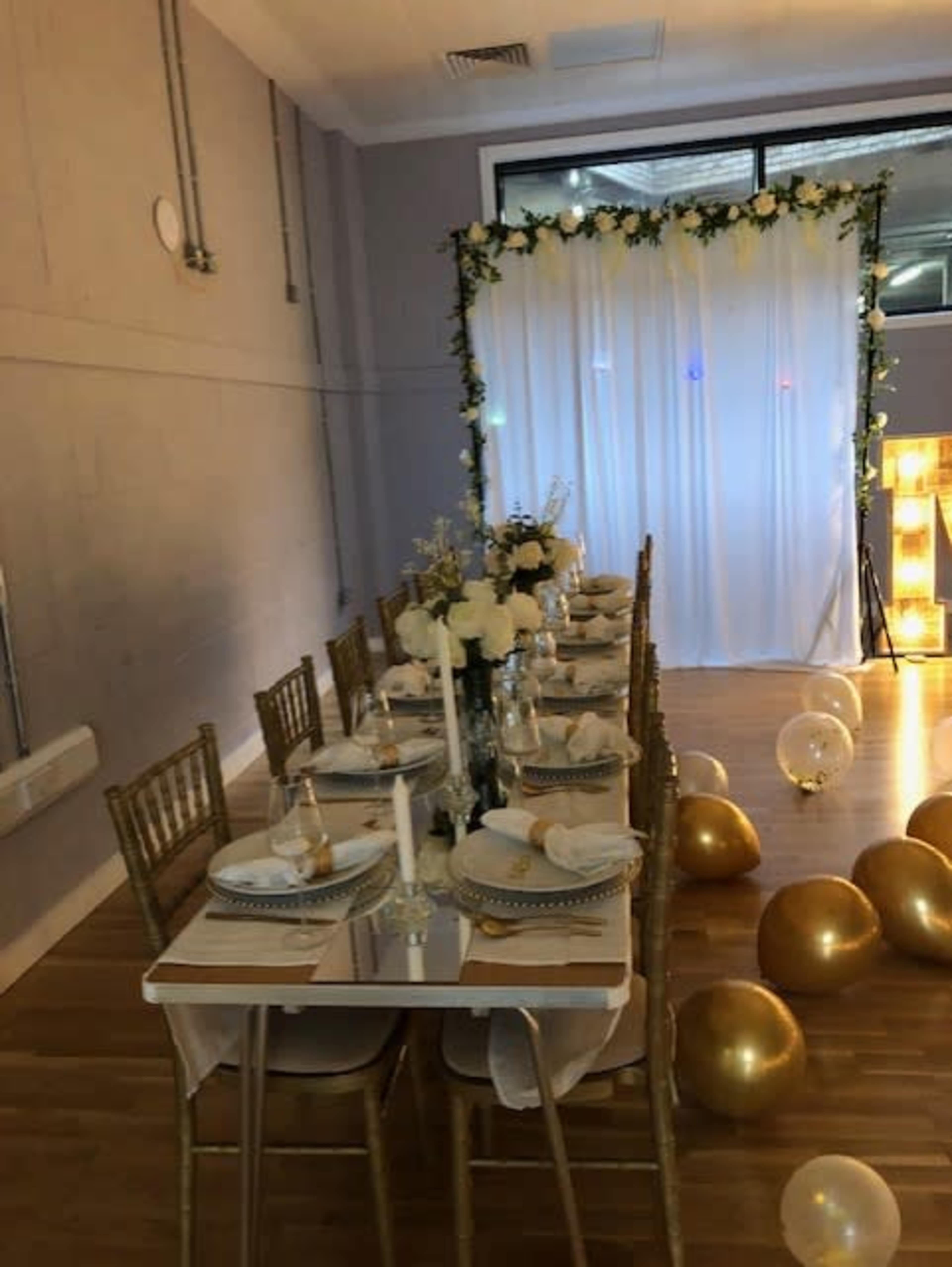 A long dining table is set with dishes and decorations in front of a backdrop of flowers and white drapery, with gold balloons scattered on the floor.