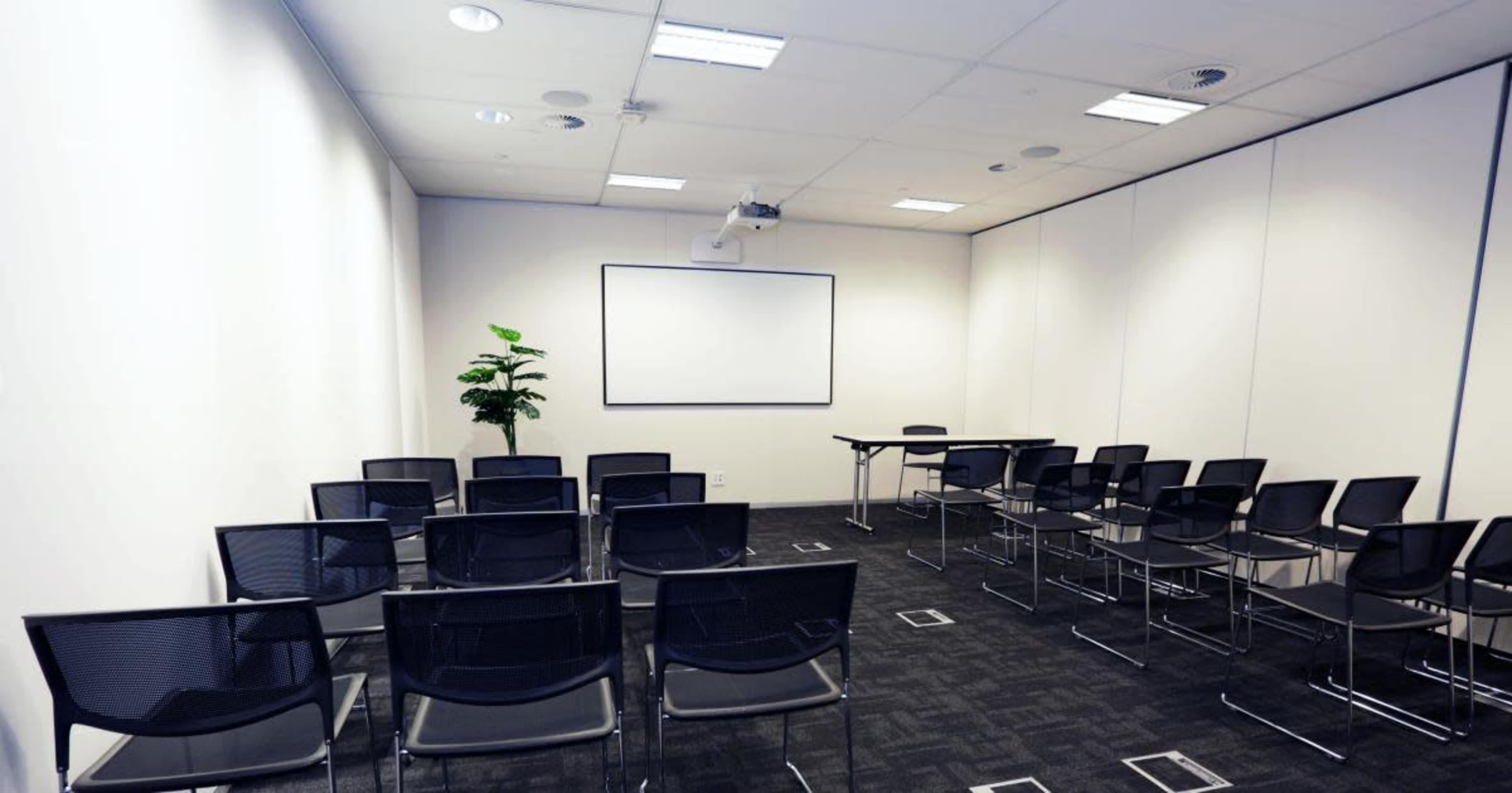 An empty meeting room contains a projector, a blank whiteboard, a table, and rows of black chairs arranged in front.