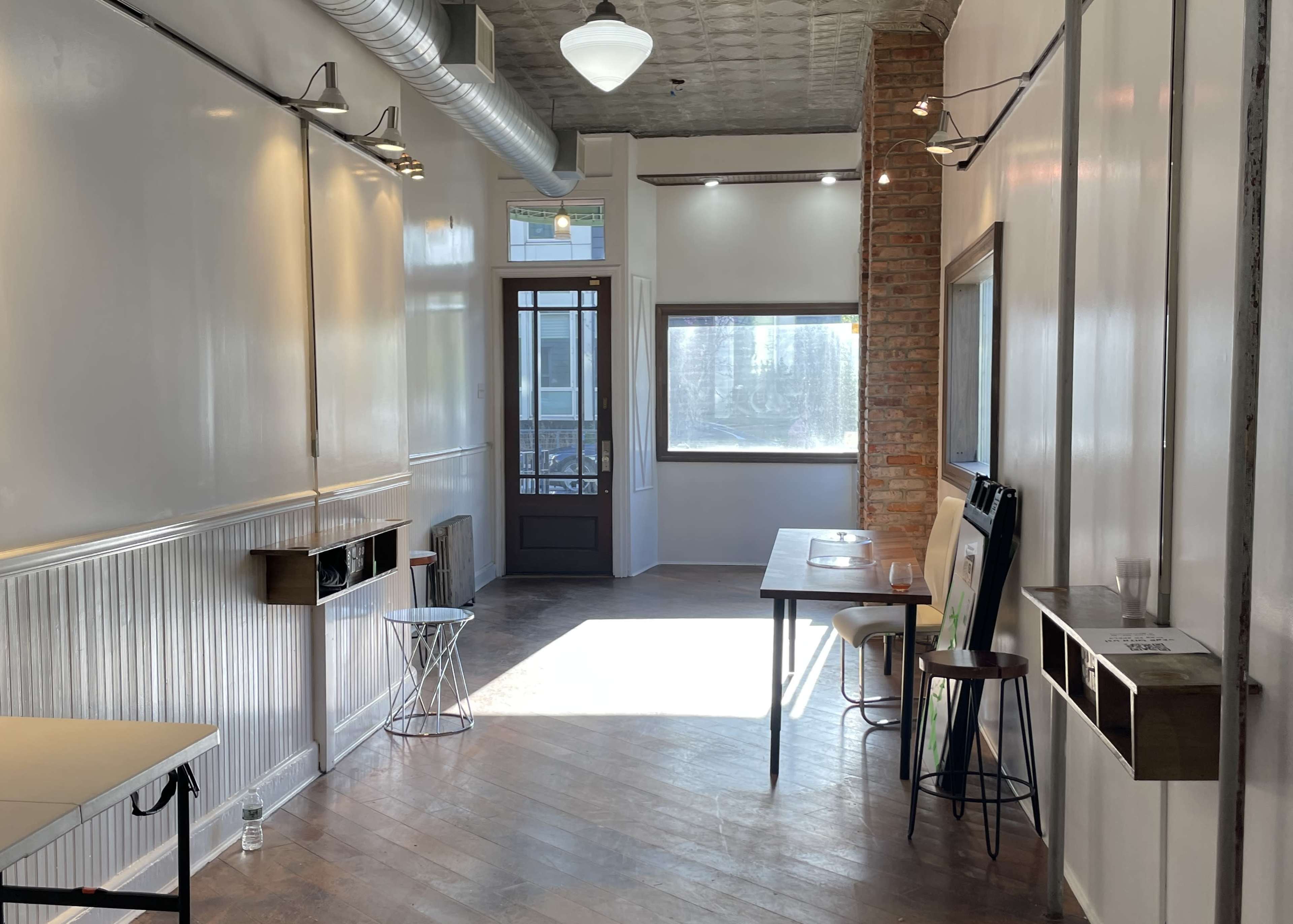 The image shows a bright, narrow hallway featuring a wooden floor, large windows, a door at one end, and tables along the sides.