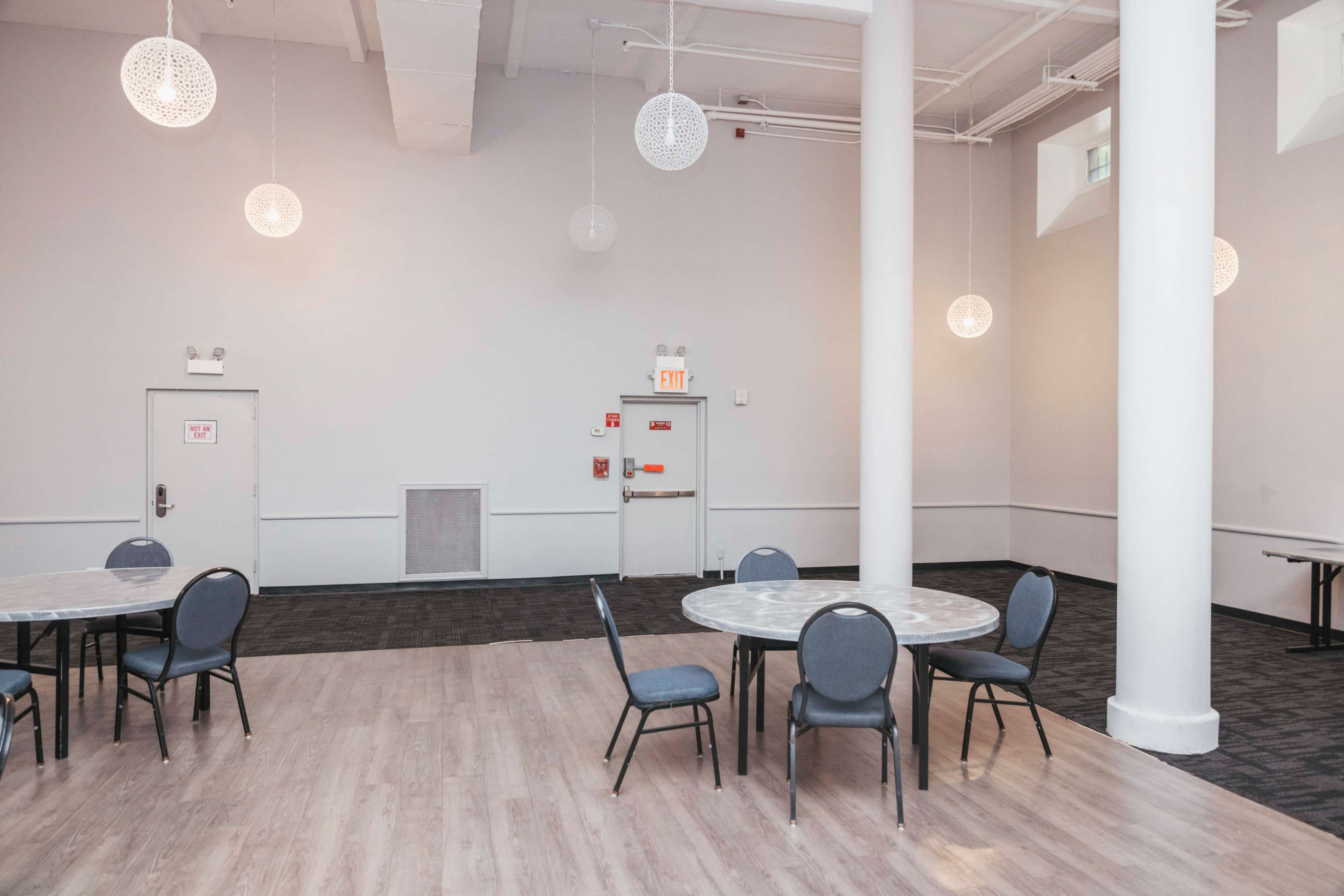 The image shows a spacious, well-lit room with several round tables and chairs arranged on a light-colored floor, featuring pendant lights and white walls.