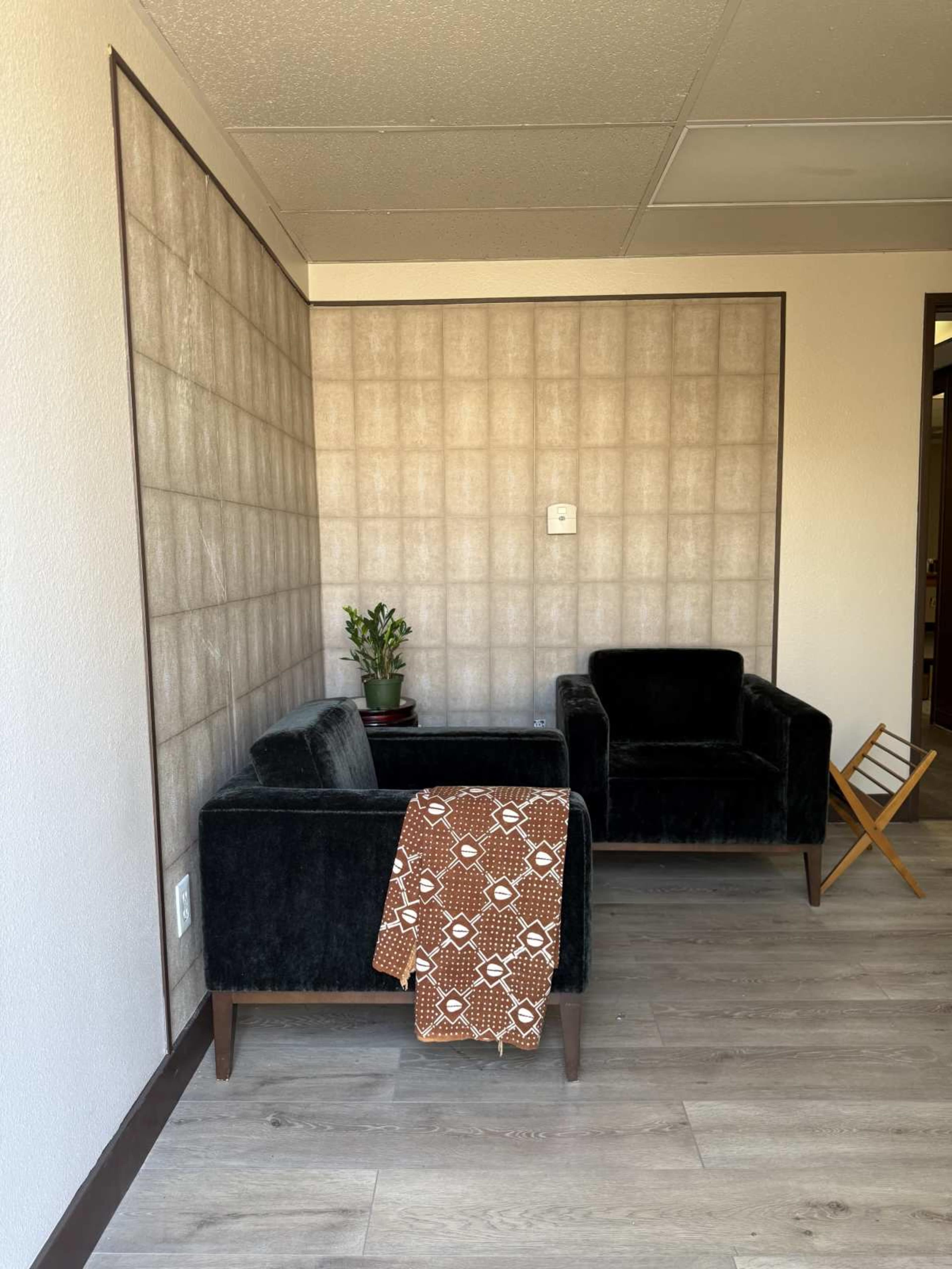The image shows a small seating area with two dark chairs, a potted plant, and a patterned blanket draped over one chair against a textured wall.