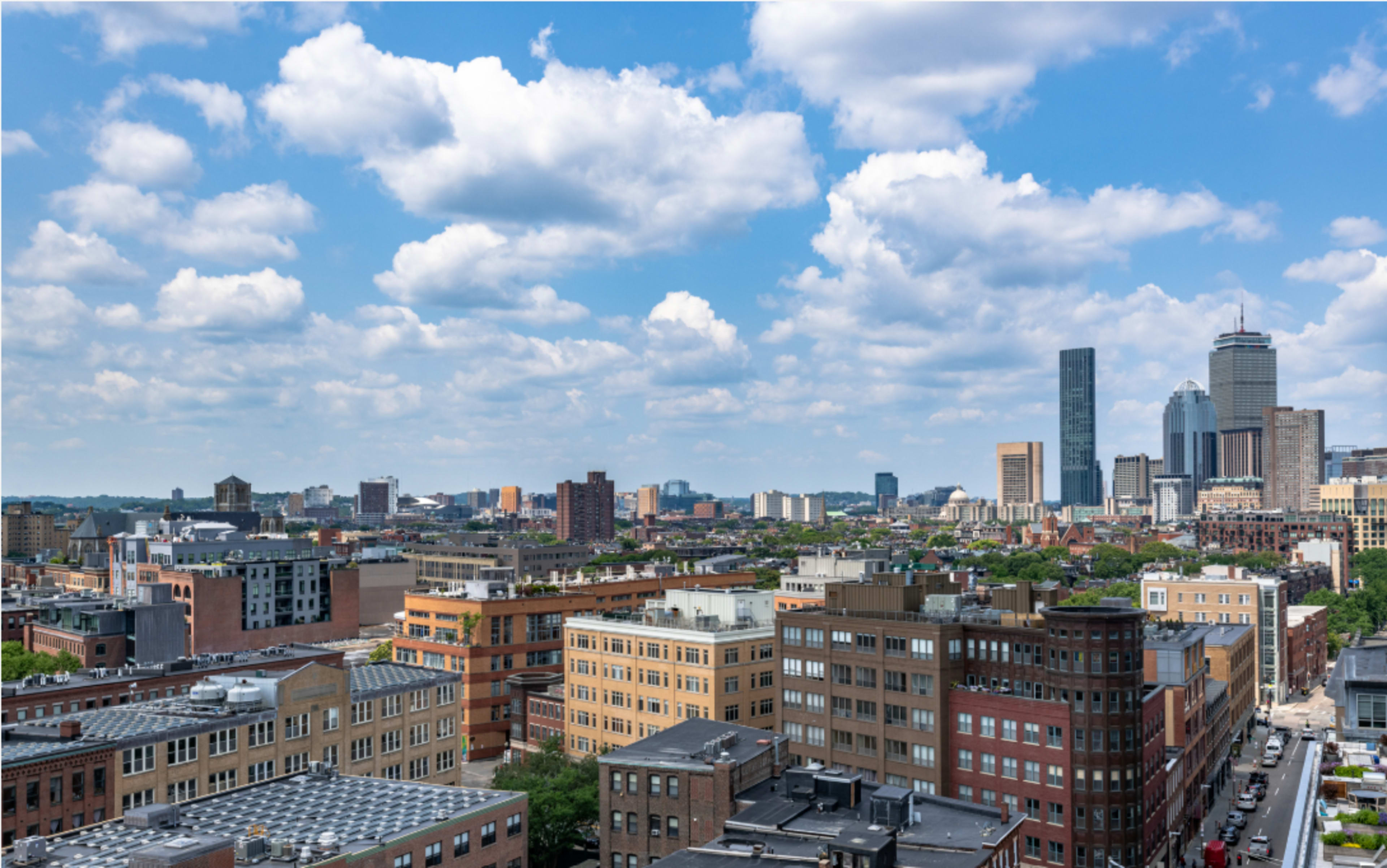 The image shows a panoramic view of a city skyline featuring a mix of residential and commercial buildings under a partly cloudy sky.