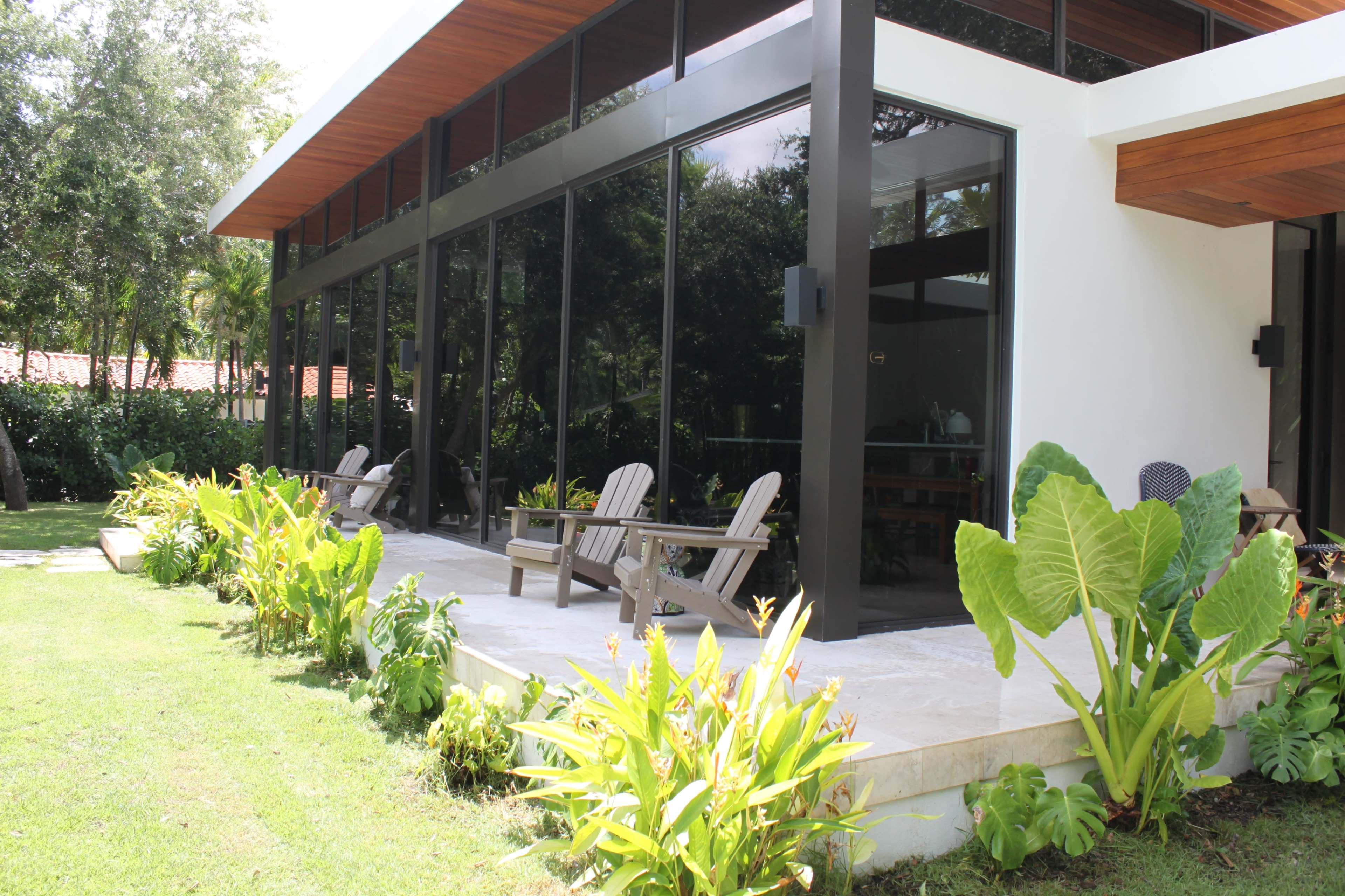 A modern house with large glass windows surrounded by tropical plants and a patio featuring wooden lounge chairs.