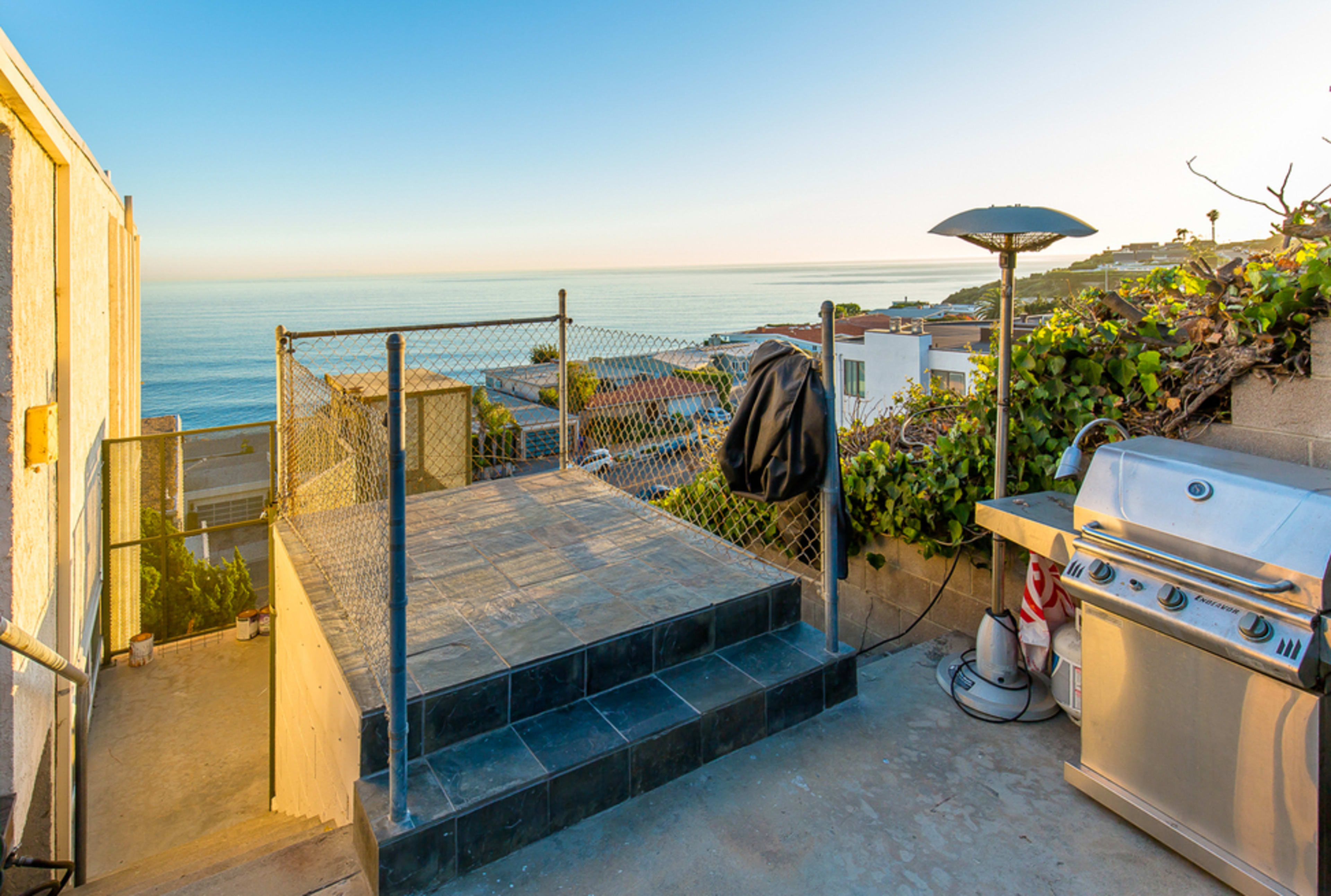 A rooftop terrace with a grill and a panoramic view of the ocean at sunset.