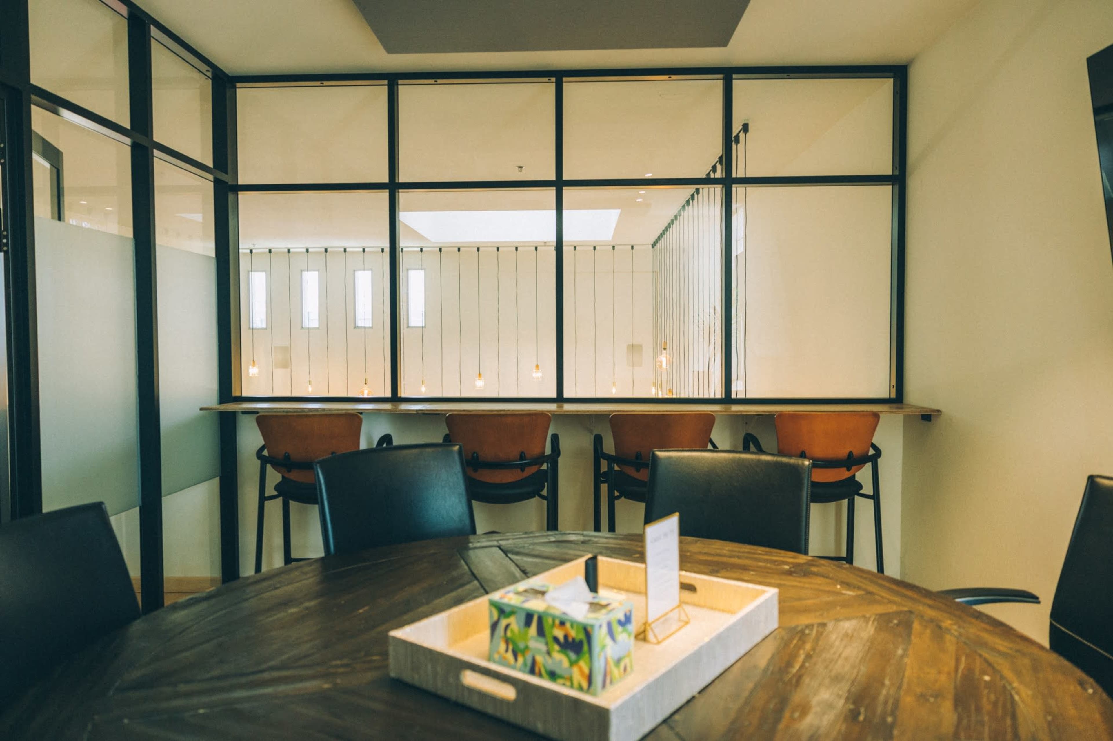 A conference room features a round wooden table in the foreground and a glass wall revealing a bar-style seating area in the background.