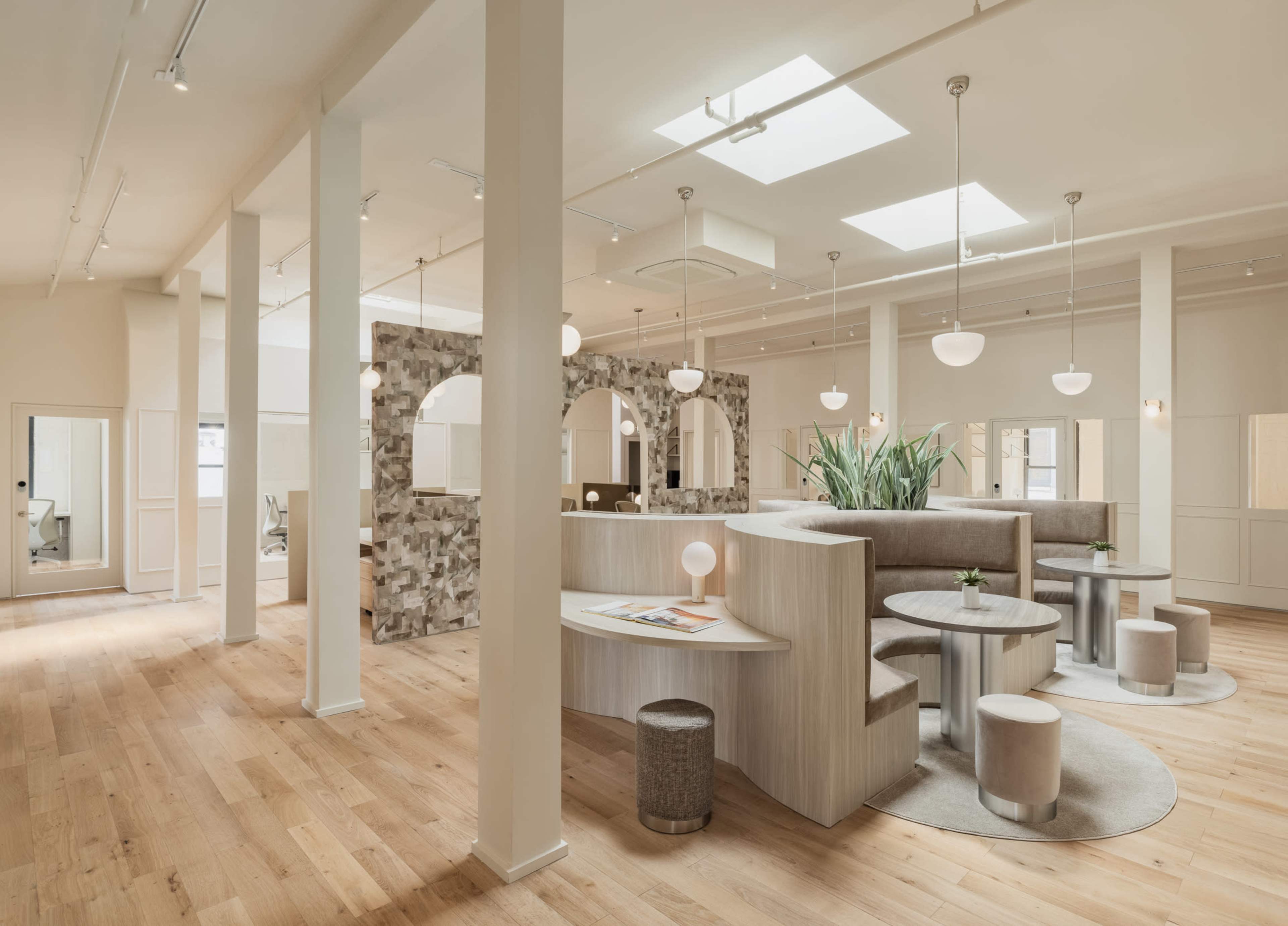 A modern, open interior space with light wooden floors, circular tables, and a combination of seating areas surrounded by neutral-colored walls and decorative elements.