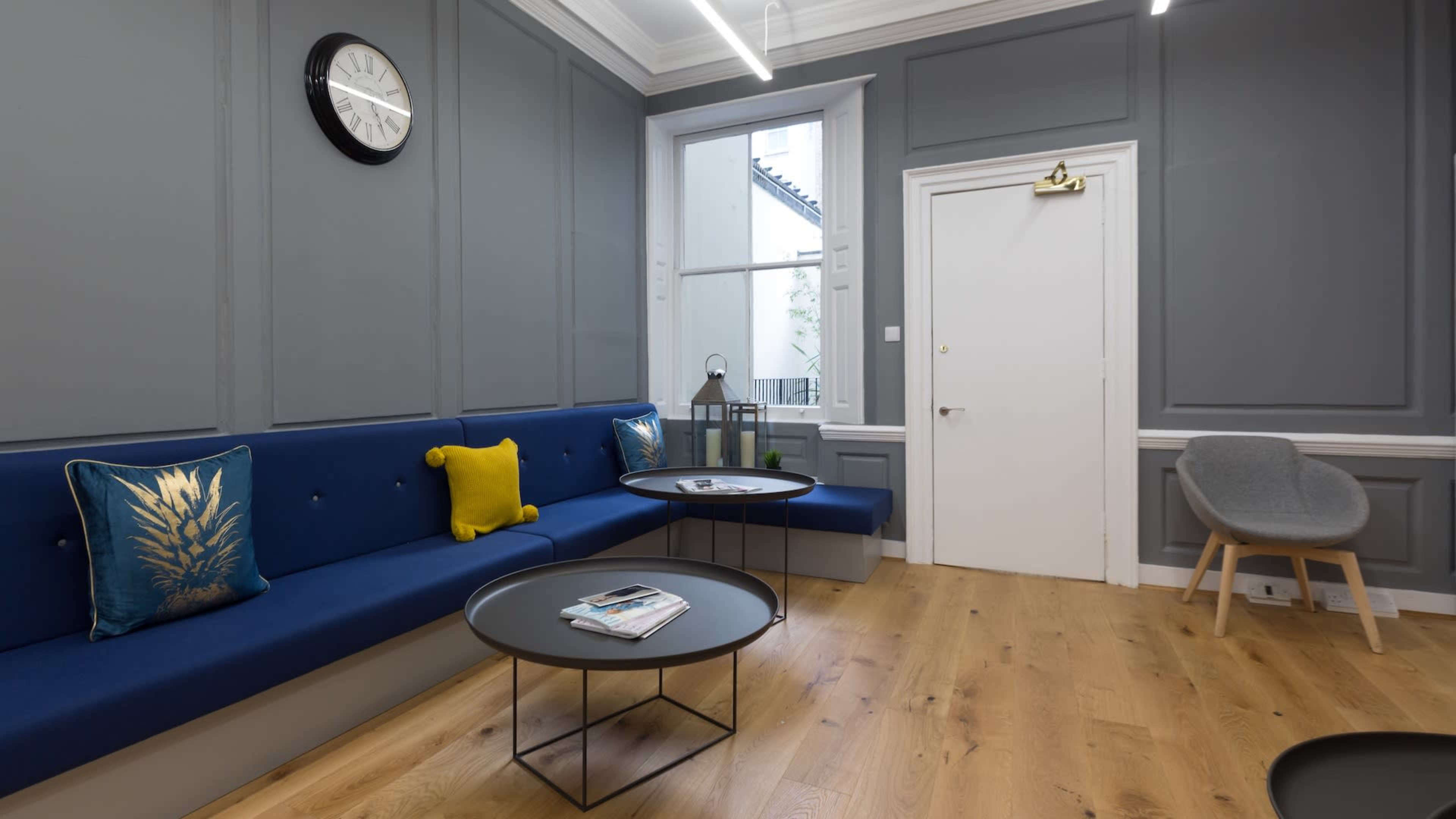 A modern waiting area featuring a blue upholstered bench, two black coffee tables, and a gray armchair, with a wall clock and a window in the background.