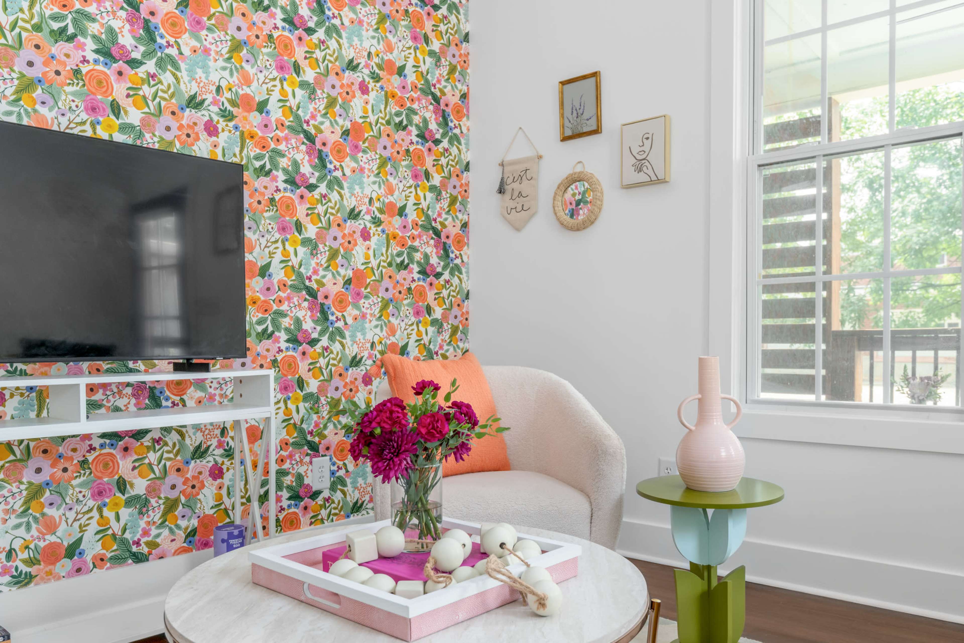 A cozy living room features a floral accent wall, a television on a white stand, a light-colored armchair with colorful cushions, and a coffee table decorated with flowers and decorative items.