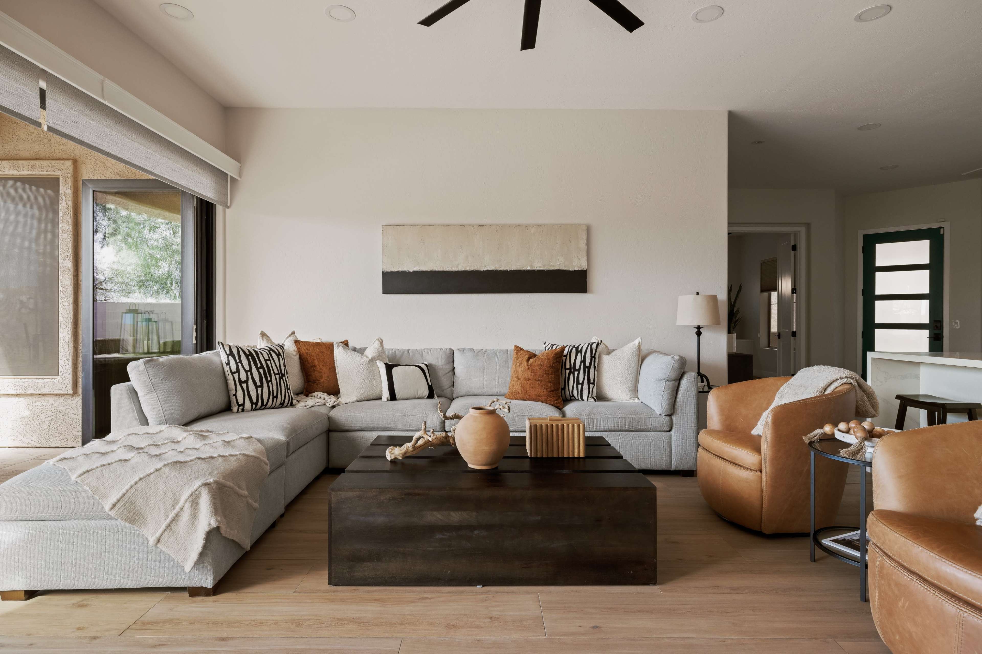 The image shows a modern living room with a large sectional sofa, a wooden coffee table, an armchair, and natural light coming through large windows.