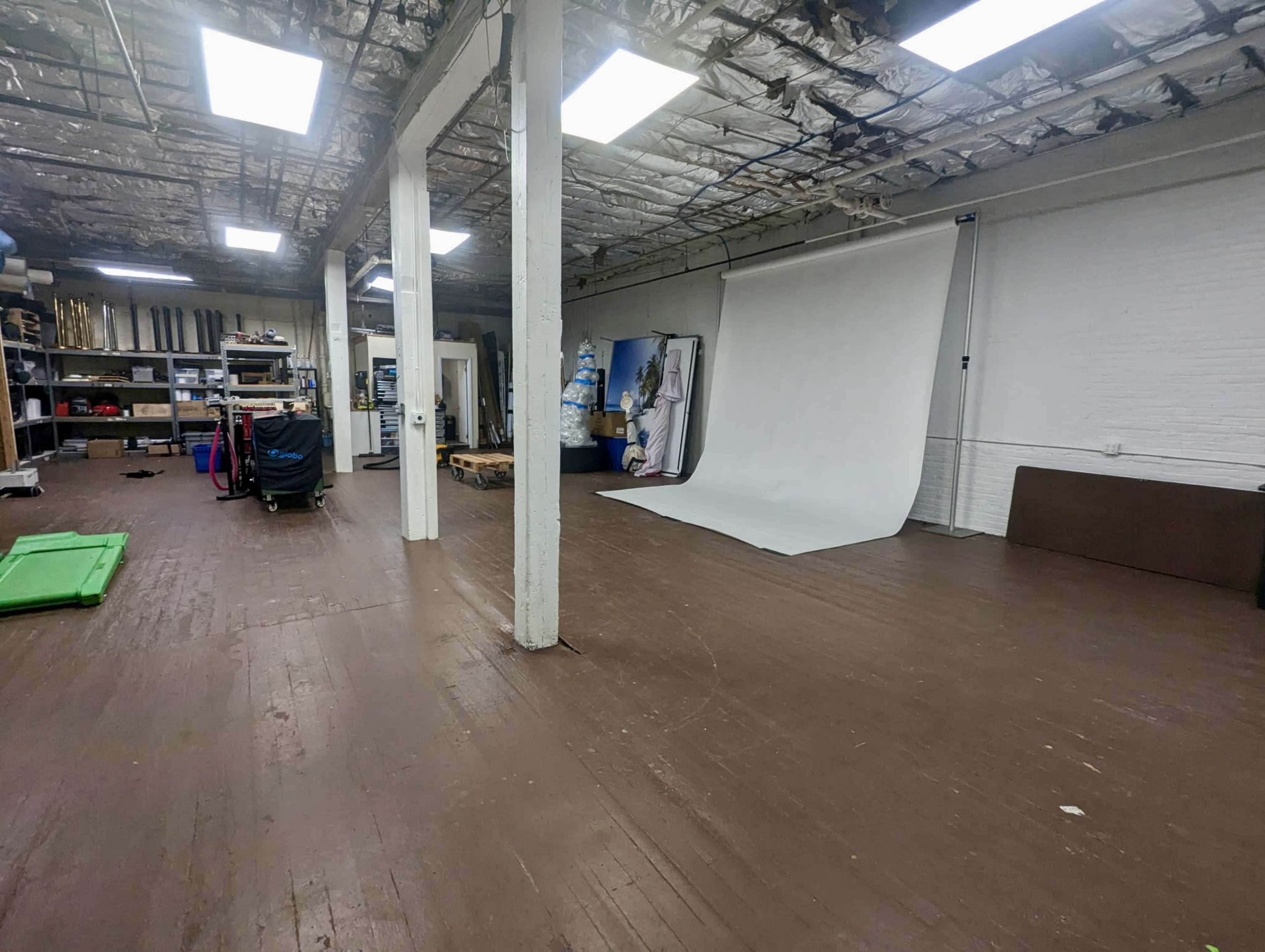 The image shows a spacious studio with wooden floors, white walls, and a large backdrop setup on the right, surrounded by various equipment and storage areas.