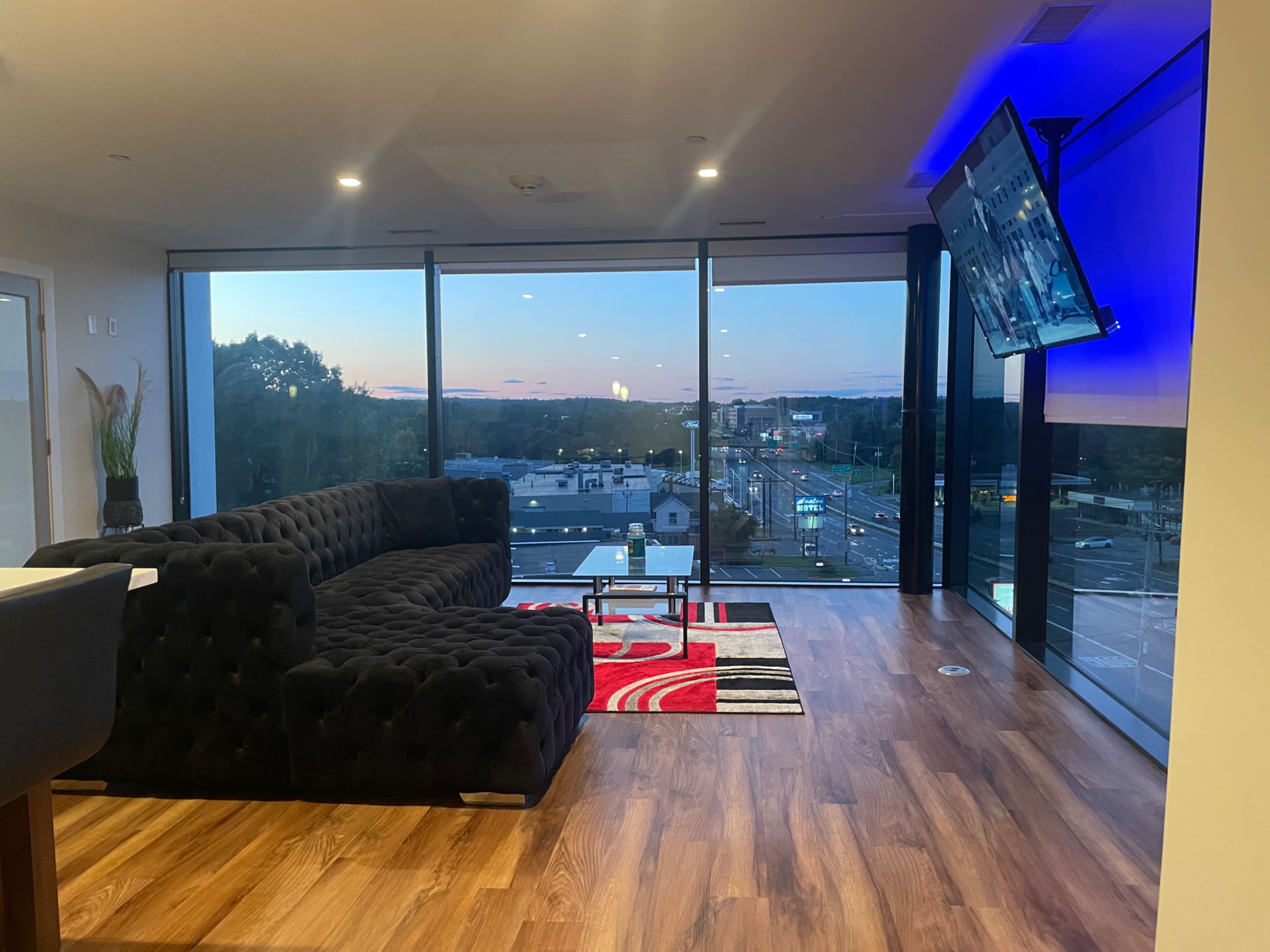 A modern living room features a black sectional sofa, a glass table, and a large TV, with floor-to-ceiling windows showcasing a sunset view over the city.