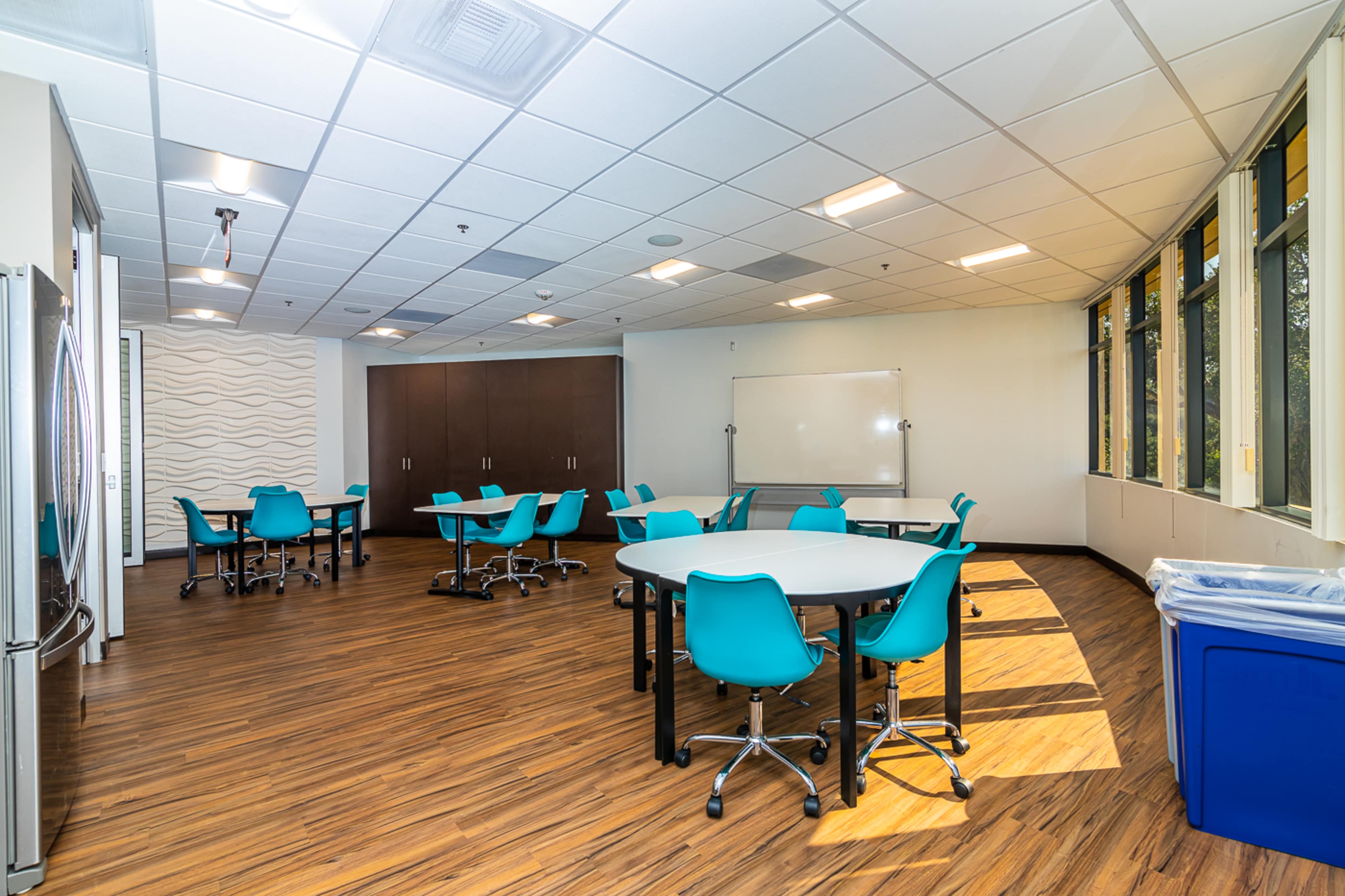 The image shows a spacious meeting room with tables and teal chairs, large windows, and a whiteboard.