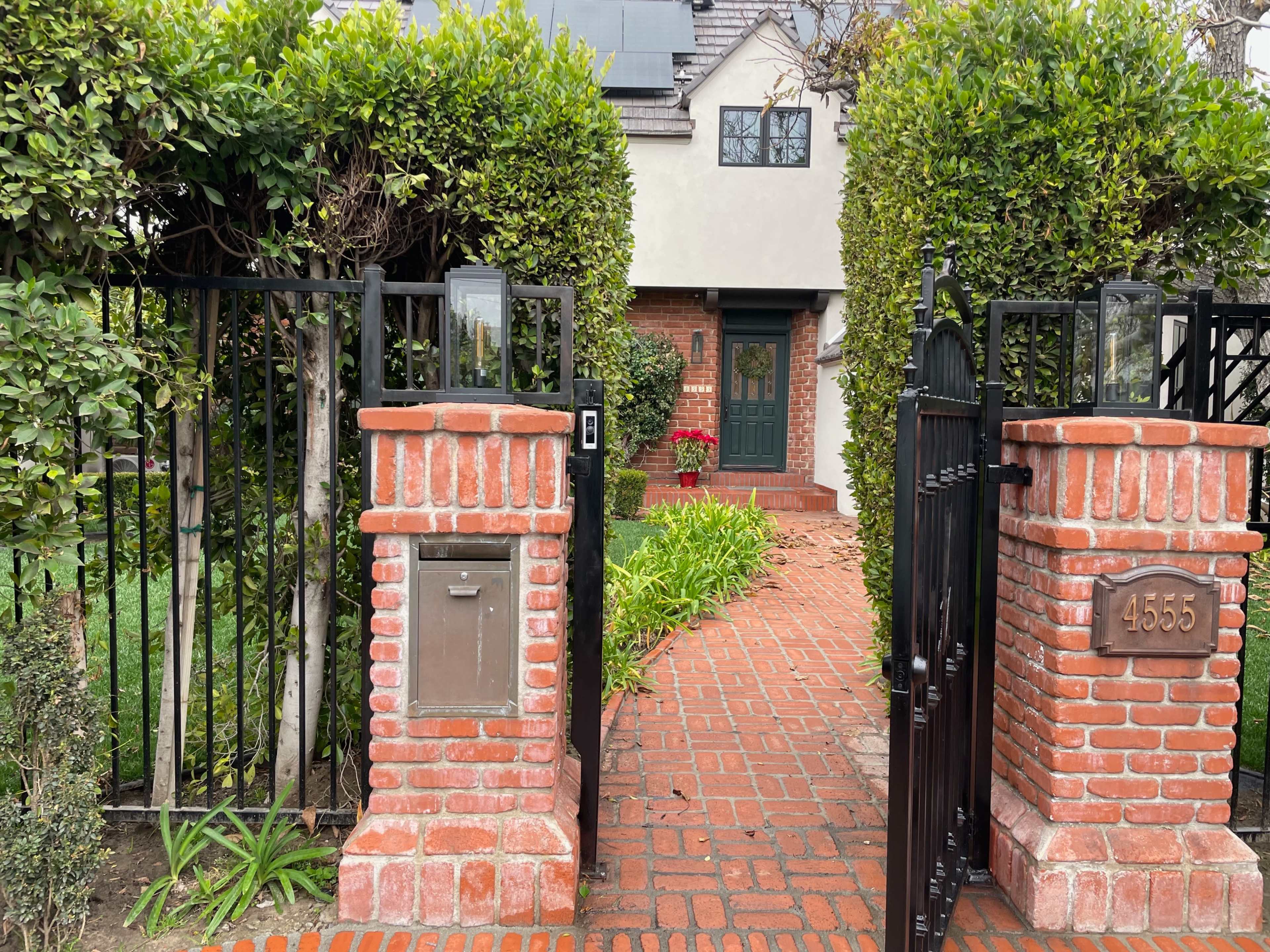 A brick pathway leading to a house, framed by two tall green hedges and a wrought-iron gate.