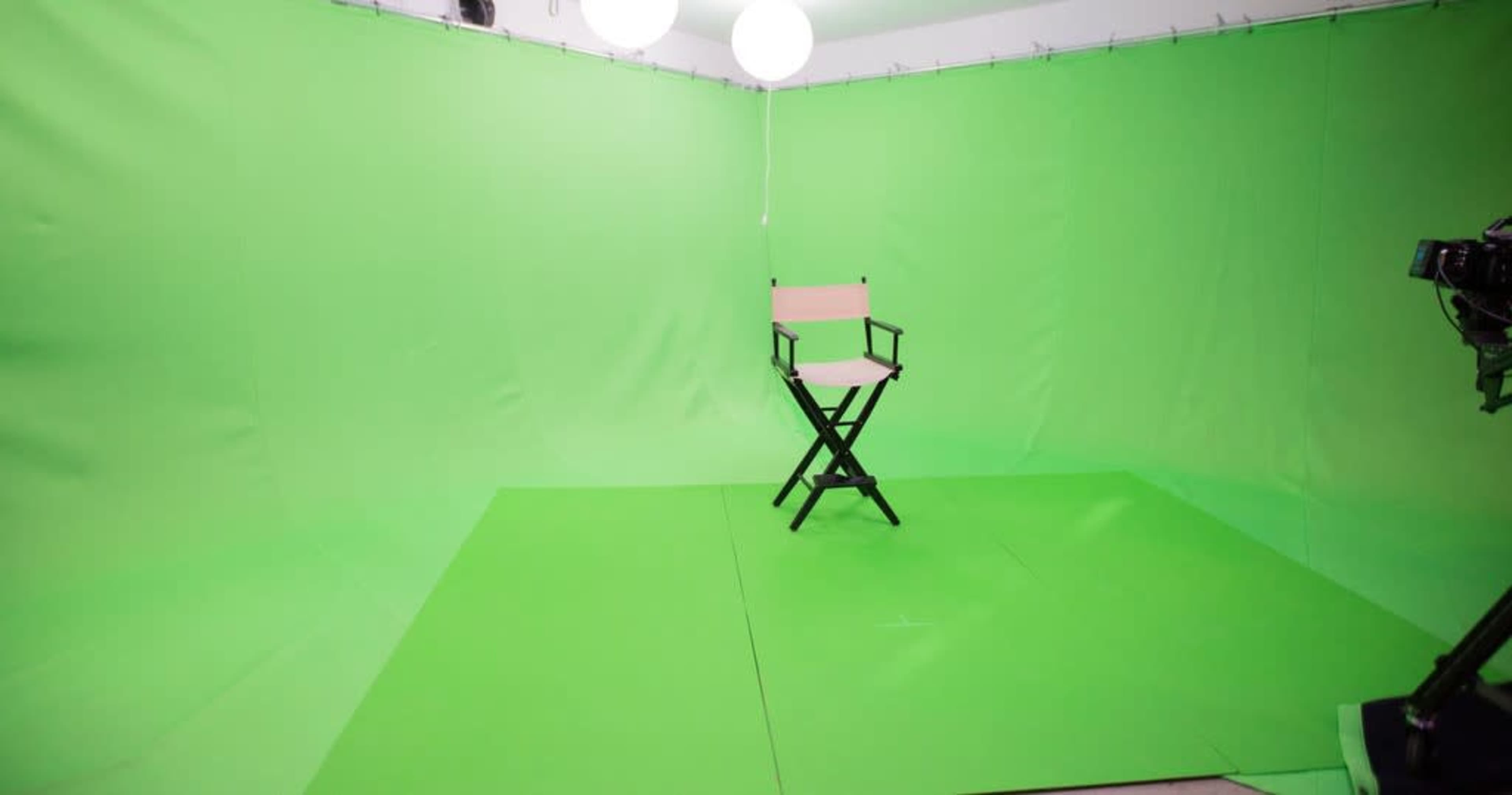 An empty green screen studio with a director's chair positioned on a green floor, illuminated by hanging lights.