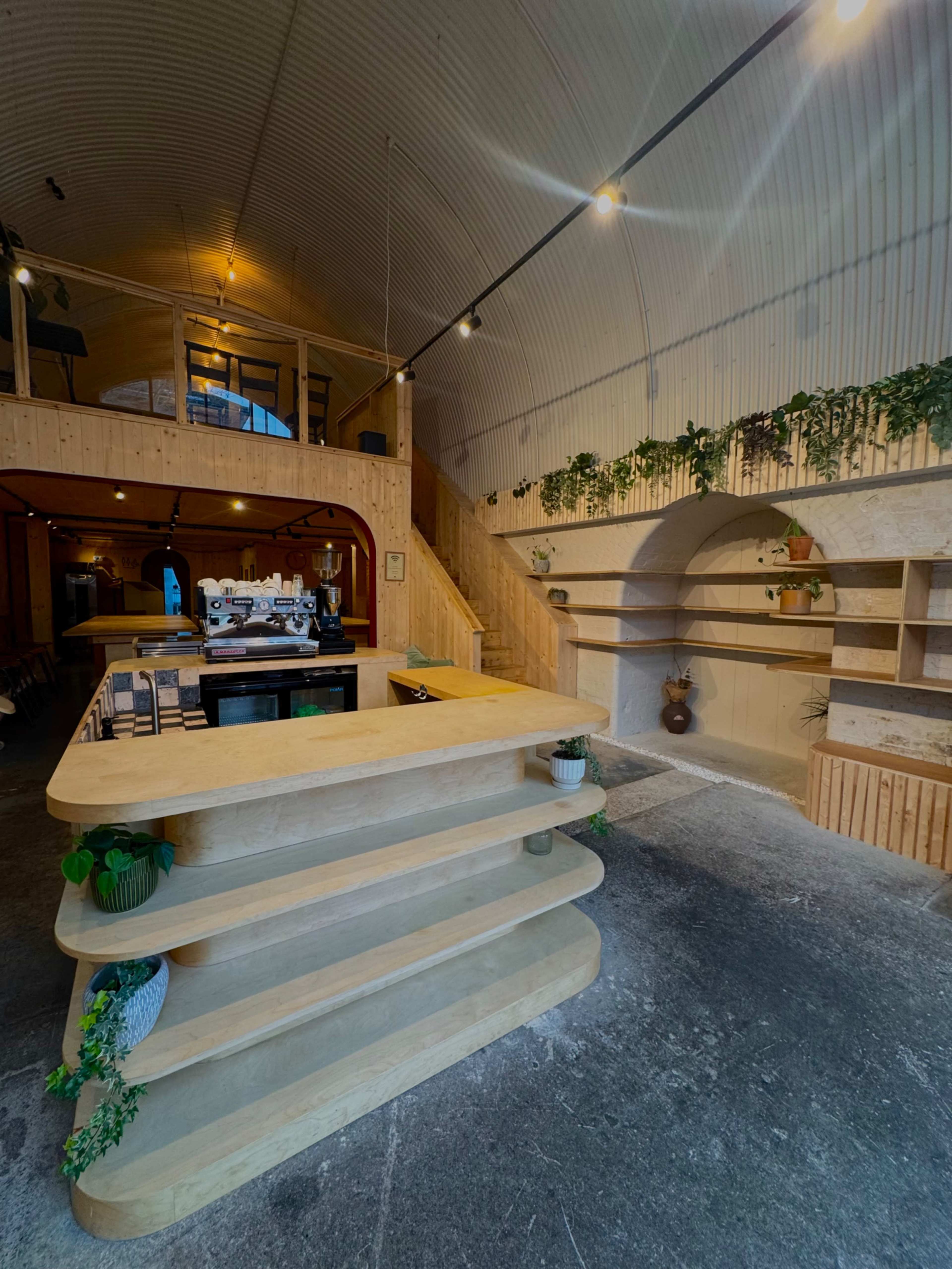 The image shows a modern, wooden café interior with a curved counter, shelves, and a staircase in the background.