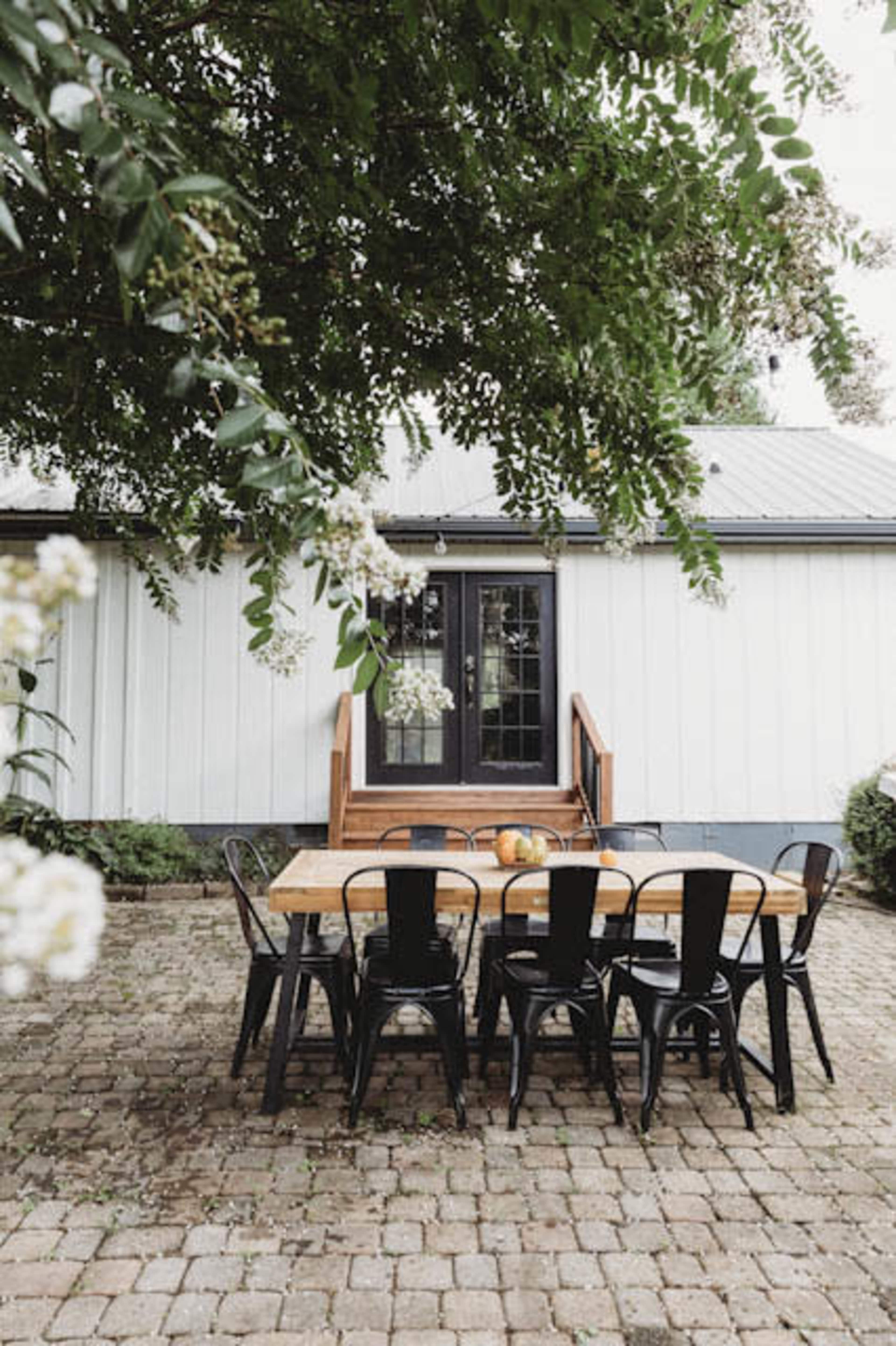 A wooden dining table is surrounded by black chairs on a brick patio outside a white and gray building.