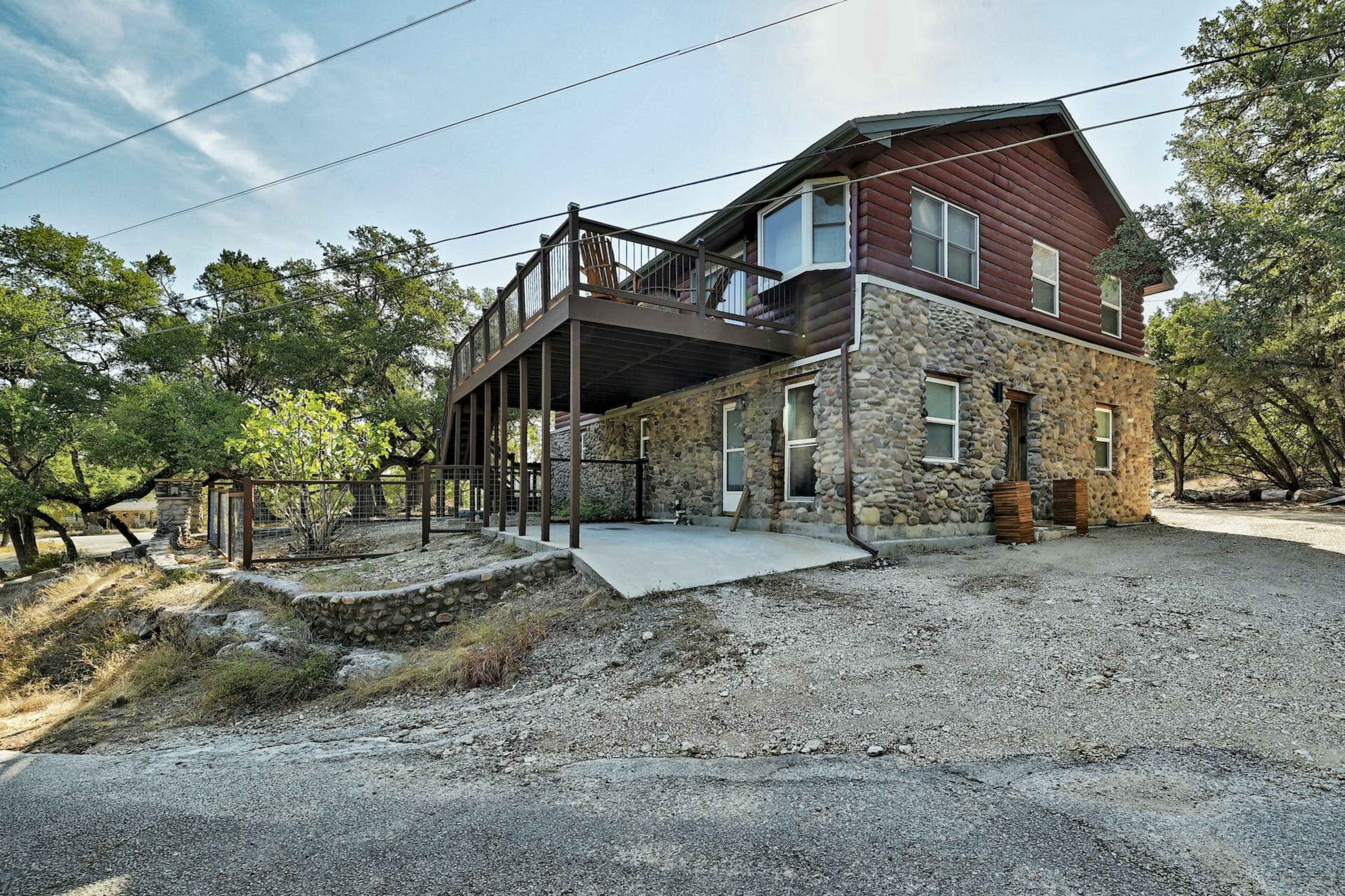 The image shows a two-story house with a stone exterior and a wooden upper deck, surrounded by trees and gravel pathways.