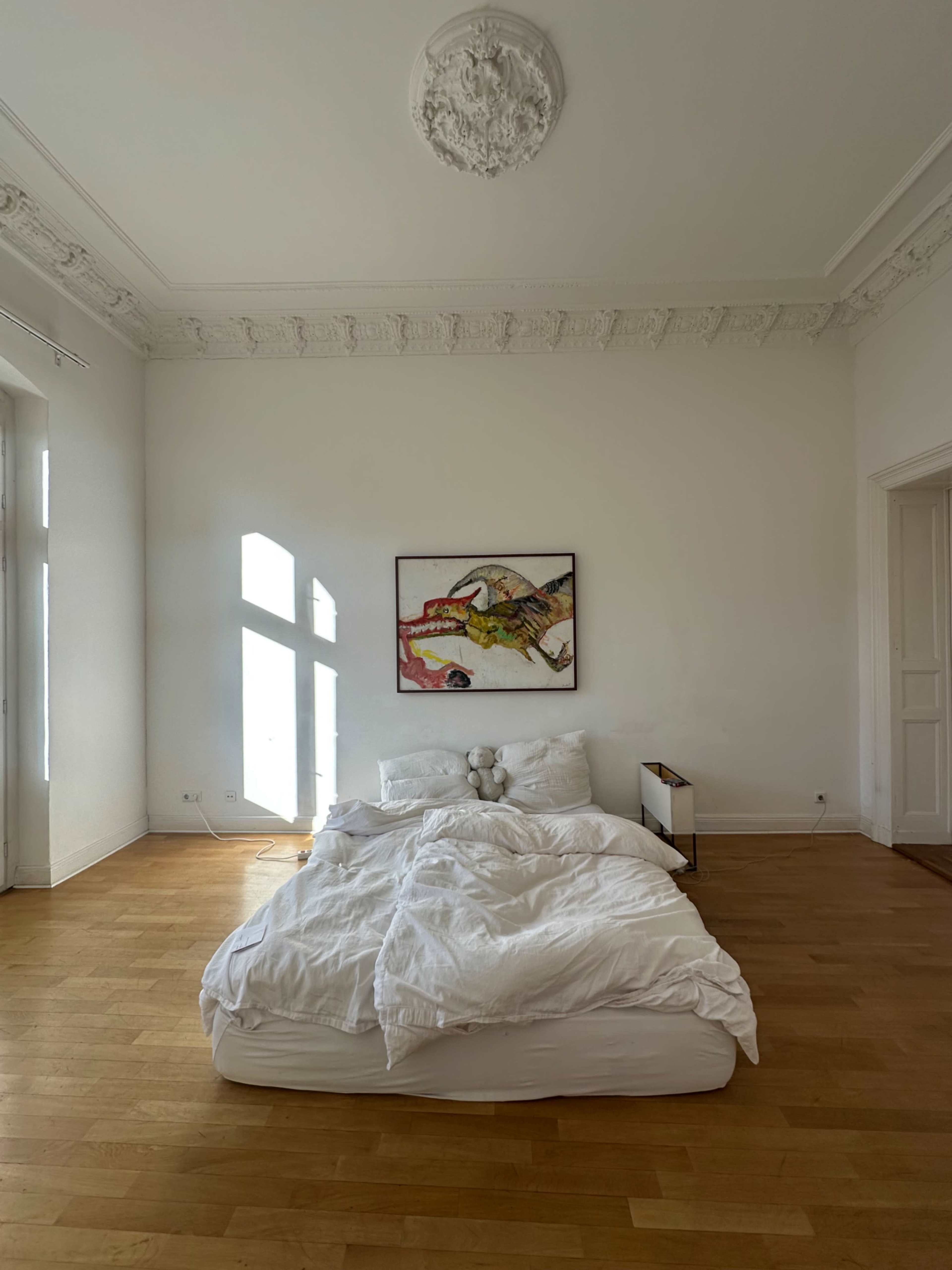 The room features a bed with white bedding, a piece of abstract art on the wall, and large windows letting in natural light.