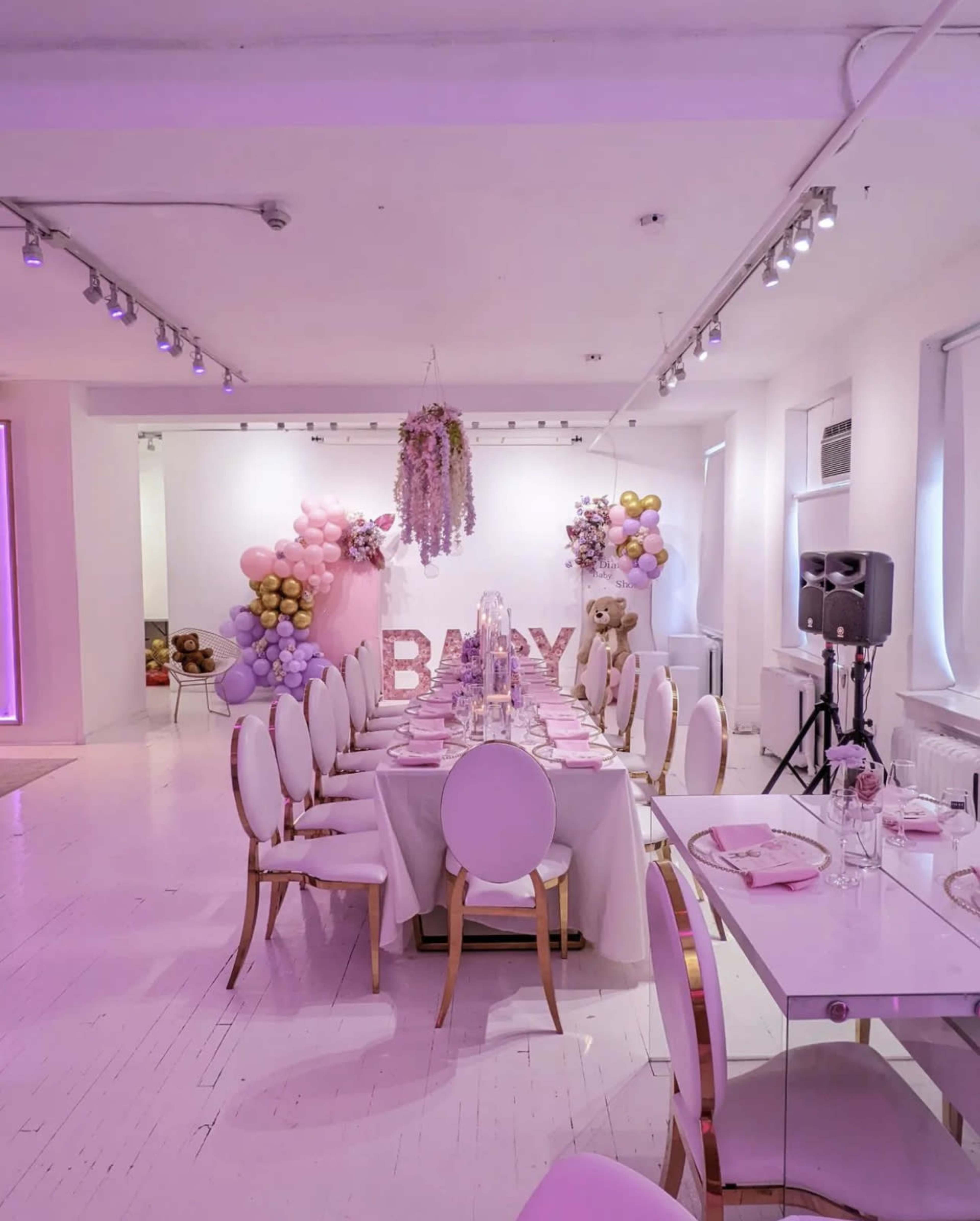 A decorated banquet room features a long table set for a celebration, surrounded by pastel balloons and floral arrangements.