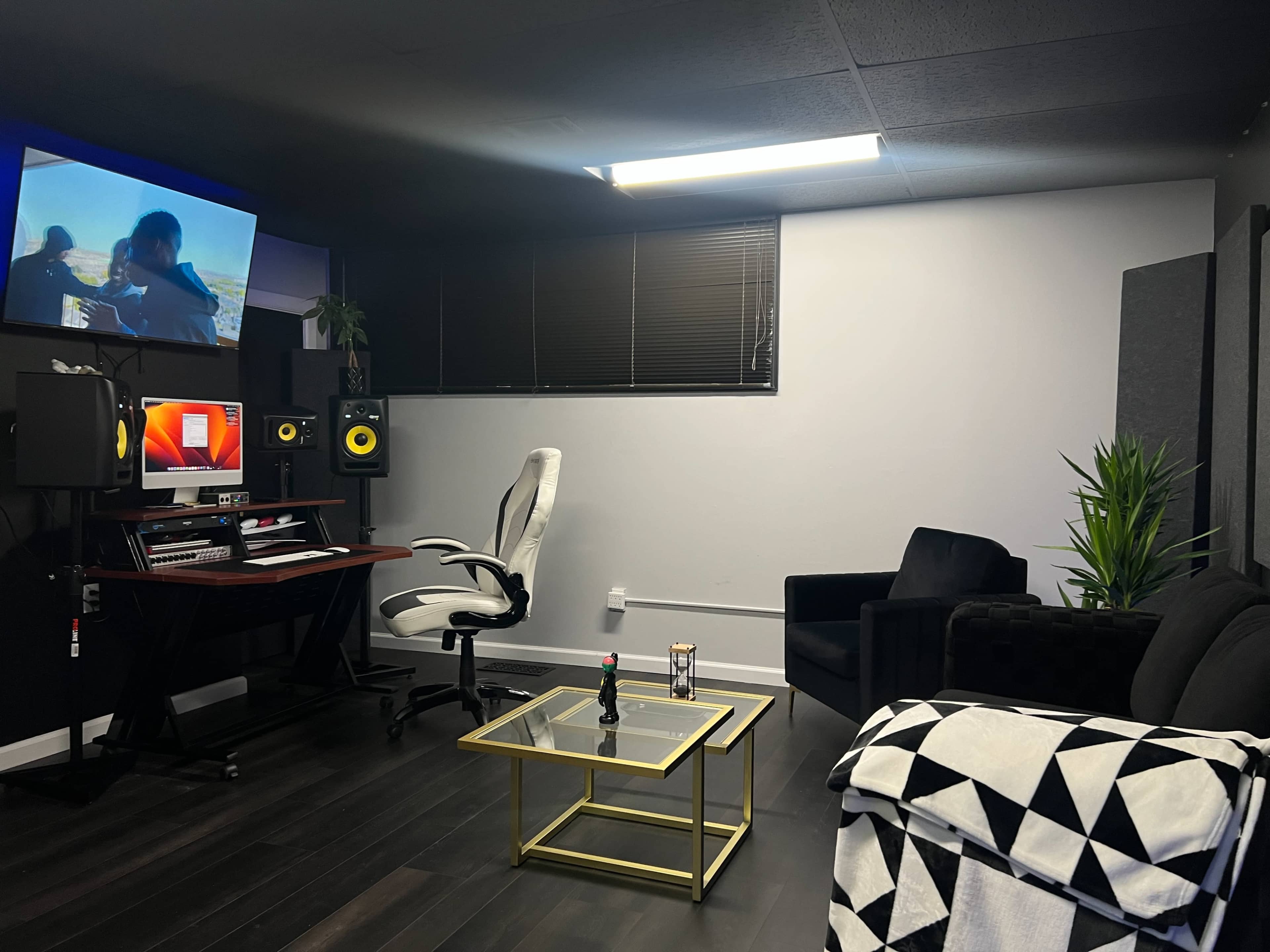 A modern music studio with a desk equipped with audio equipment, a comfortable chair, a couch, a golden coffee table, and a wall-mounted screen displaying visuals.