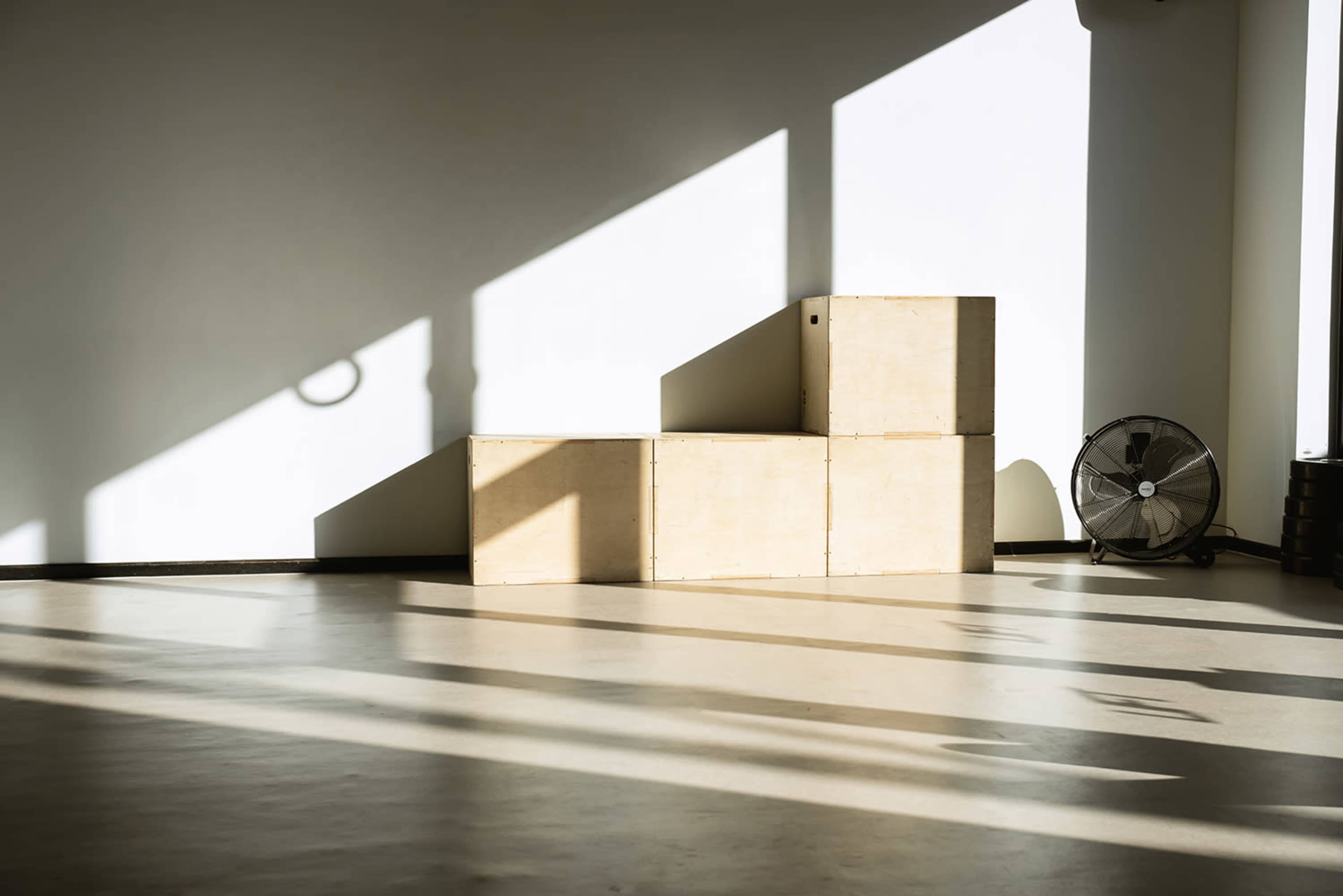 Three wooden boxes arranged in a stair-like formation in a well-lit room, with a fan positioned nearby and shadows cast on the floor.
