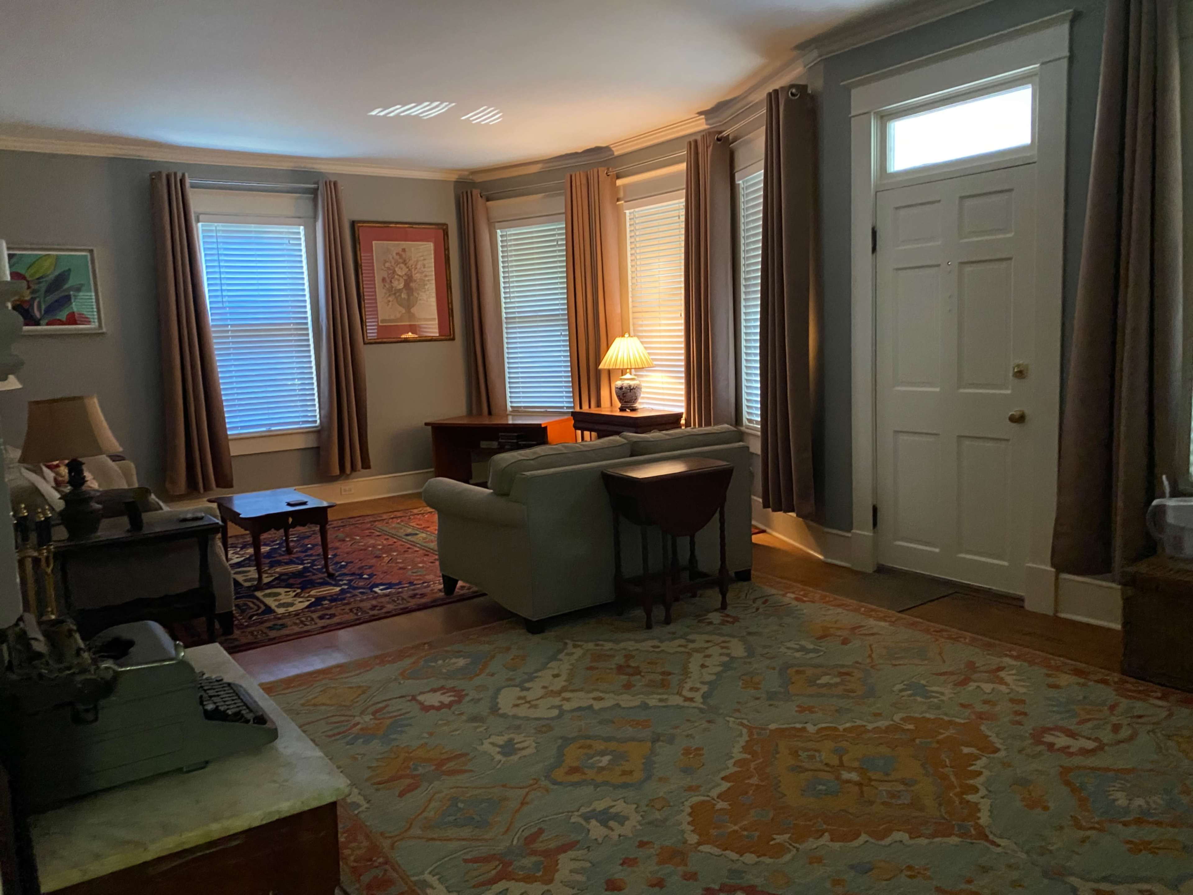 The room features gray walls, a seating area with a sofa and chairs, a wooden table, and prominent sunlight coming through the windows.