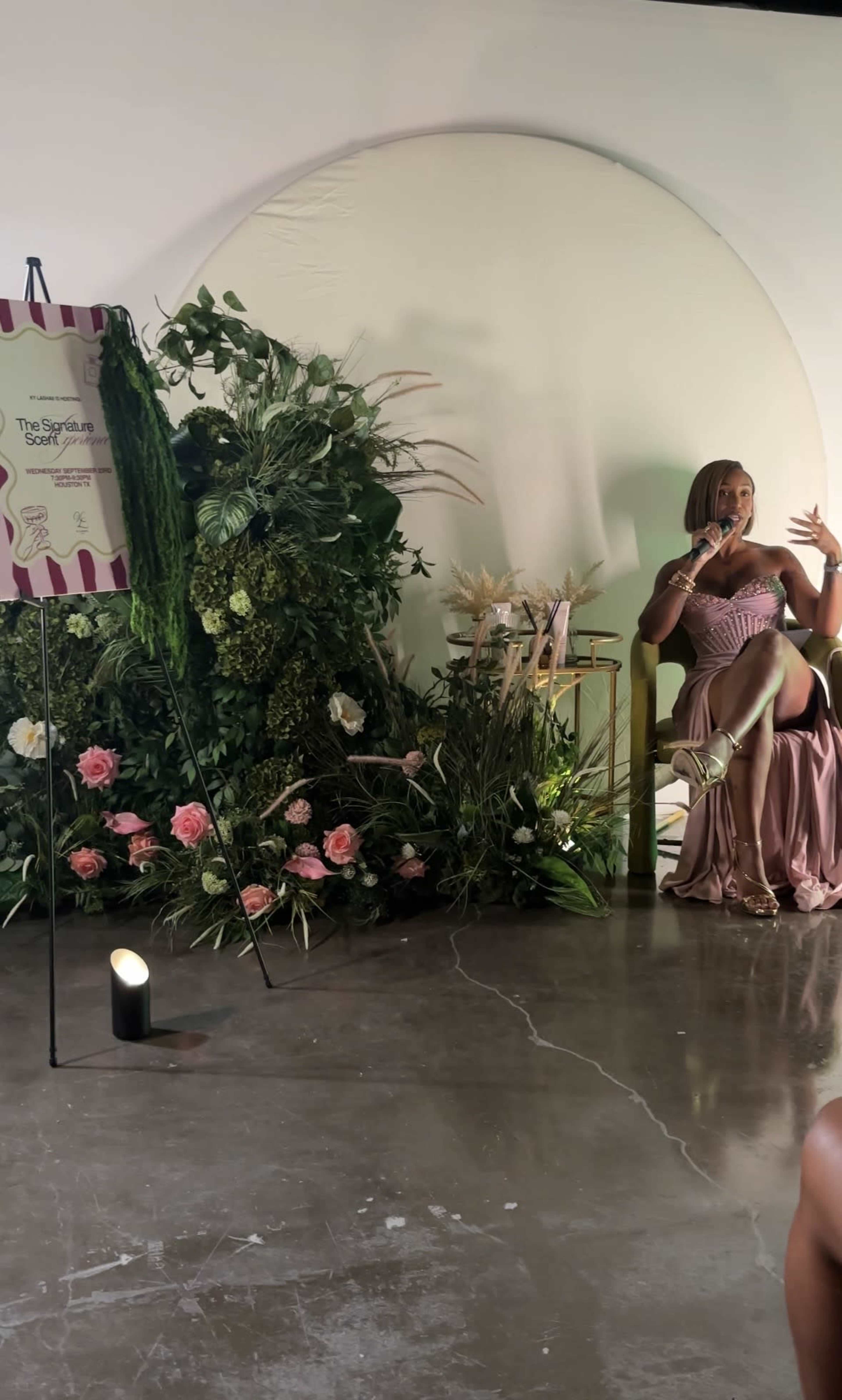 A woman in a pink dress sits on a chair in front of a decorative backdrop featuring greenery and flowers, with a sign nearby.